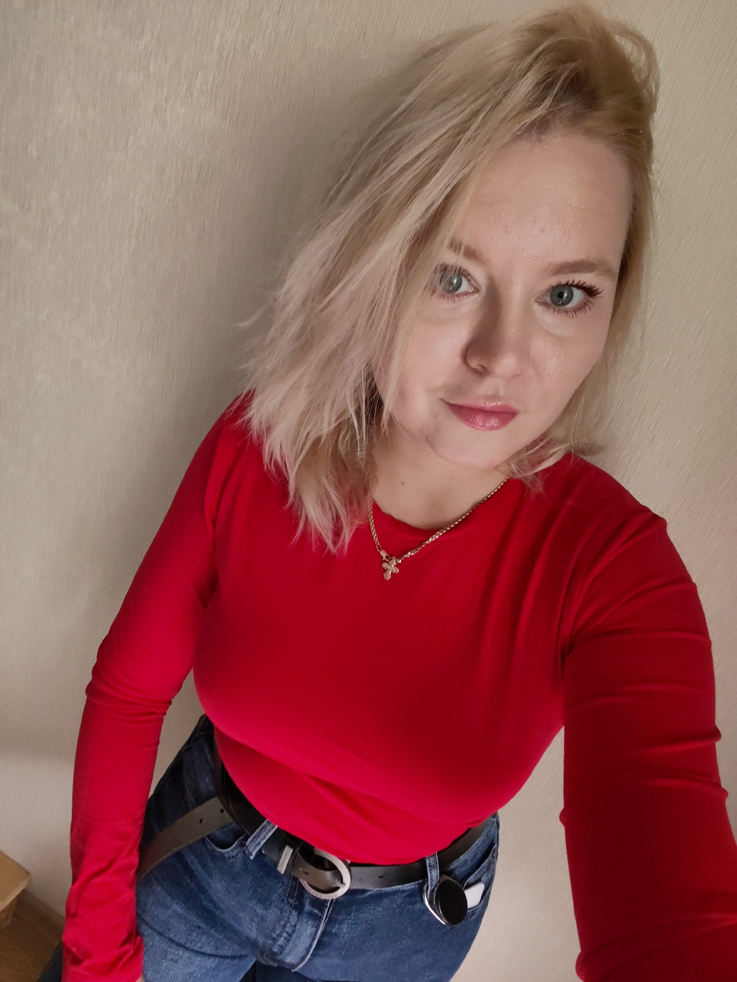 In this high-angle, indoor selfie, a woman with blonde hair and light blue eyes poses against a neutral-colored wall. She is wearing a red long-sleeved shirt and blue jeans, accessorized with a dark belt and a delicate gold necklace with a small pendant. The woman is looking directly at the camera with a slight smile. The environment appears to be a domestic setting. The photograph was taken in Izhevsk, Russia.