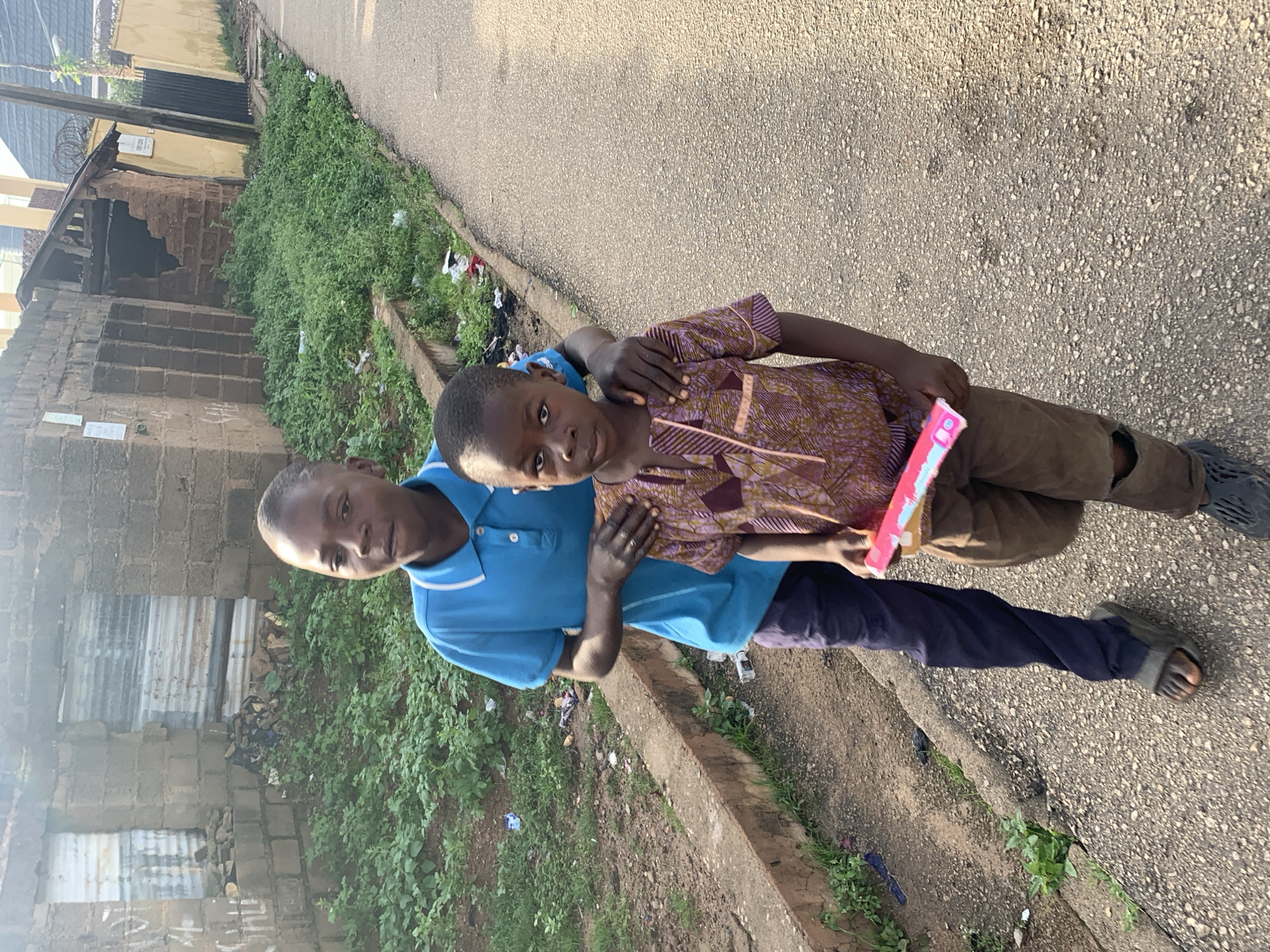 Two young boys are walking on a paved street in Jos, Nigeria. The boy in front, wearing a patterned brown shirt and torn brown pants, is looking at the camera. The boy behind him, wearing a blue polo shirt and dark pants, has his hand on the other boy's shoulder. The boy in front is holding a pink and blue object, possibly a toy. The background shows a neglected building with overgrown vegetation and debris. The lighting suggests it is late afternoon.