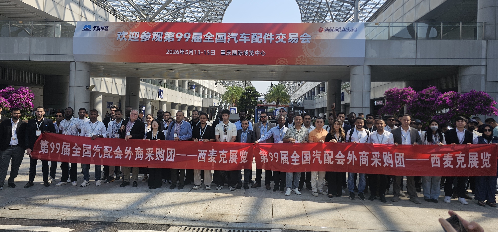 A large group of men and women are gathered outdoors, holding a red banner that reads "The 99th National Auto Parts Fair Foreign Procurement Group - Cimac Exhibition, The 99th National Auto Parts Fair Foreign Procurement Group - Cimac Exhibition". Above them, another banner states "Welcome to visit the 99th National Auto Parts Fair, May 13-15, 2026, Chongqing International Expo Center". The logo of SINOMACHINT is visible on the upper left of the banner. The group stands in front of a modern building with an elaborate glass and steel roof structure. Purple flowering bushes are situated on either side of the group. The scene is well-lit and appears to be located in Liangjiang, China, given the event location.