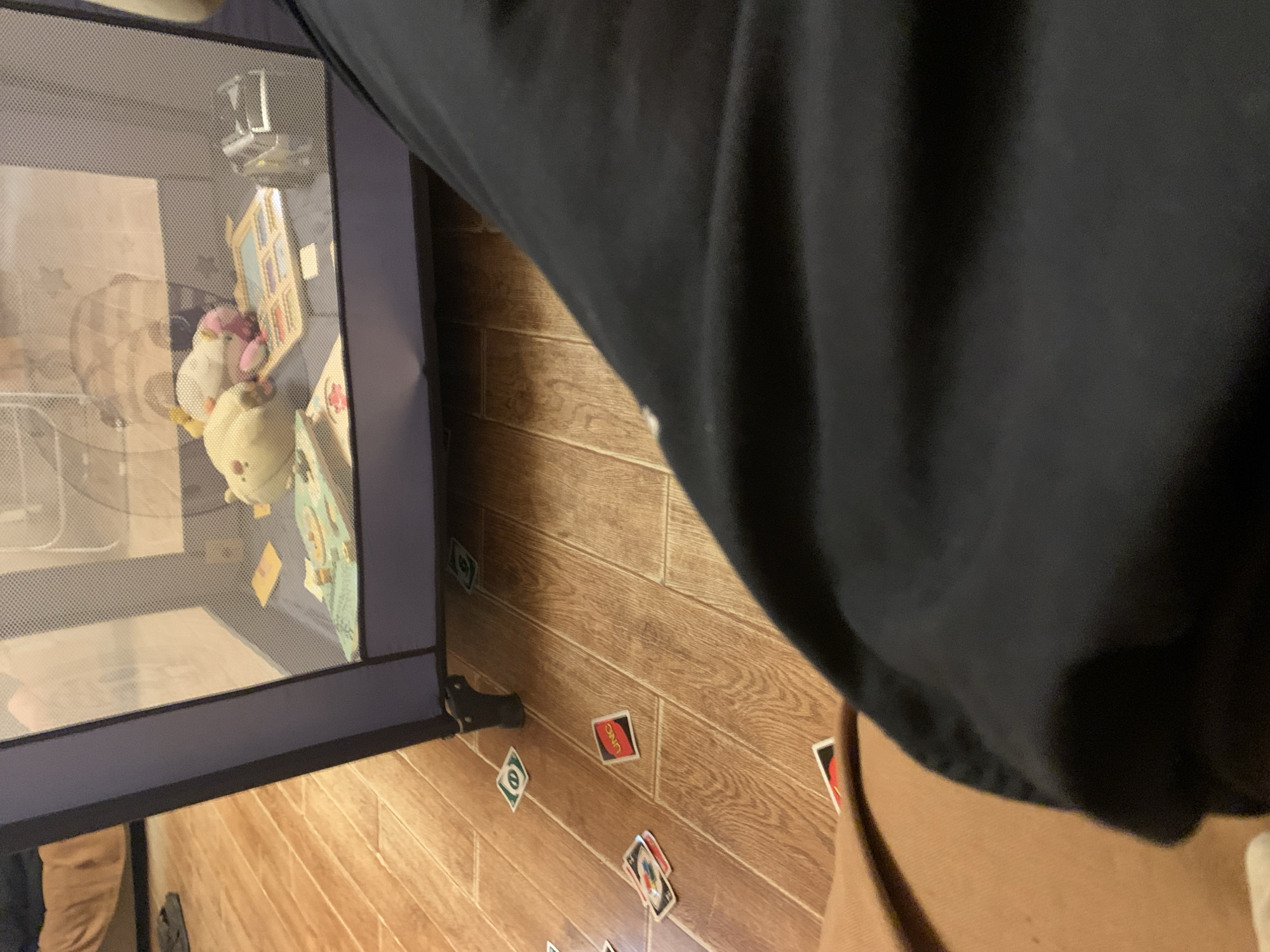 The image shows a close-up of a playpen filled with stuffed animals and a children's toy. Uno cards are scattered on the wooden floor surrounding the playpen. The scene appears to be in a home environment.