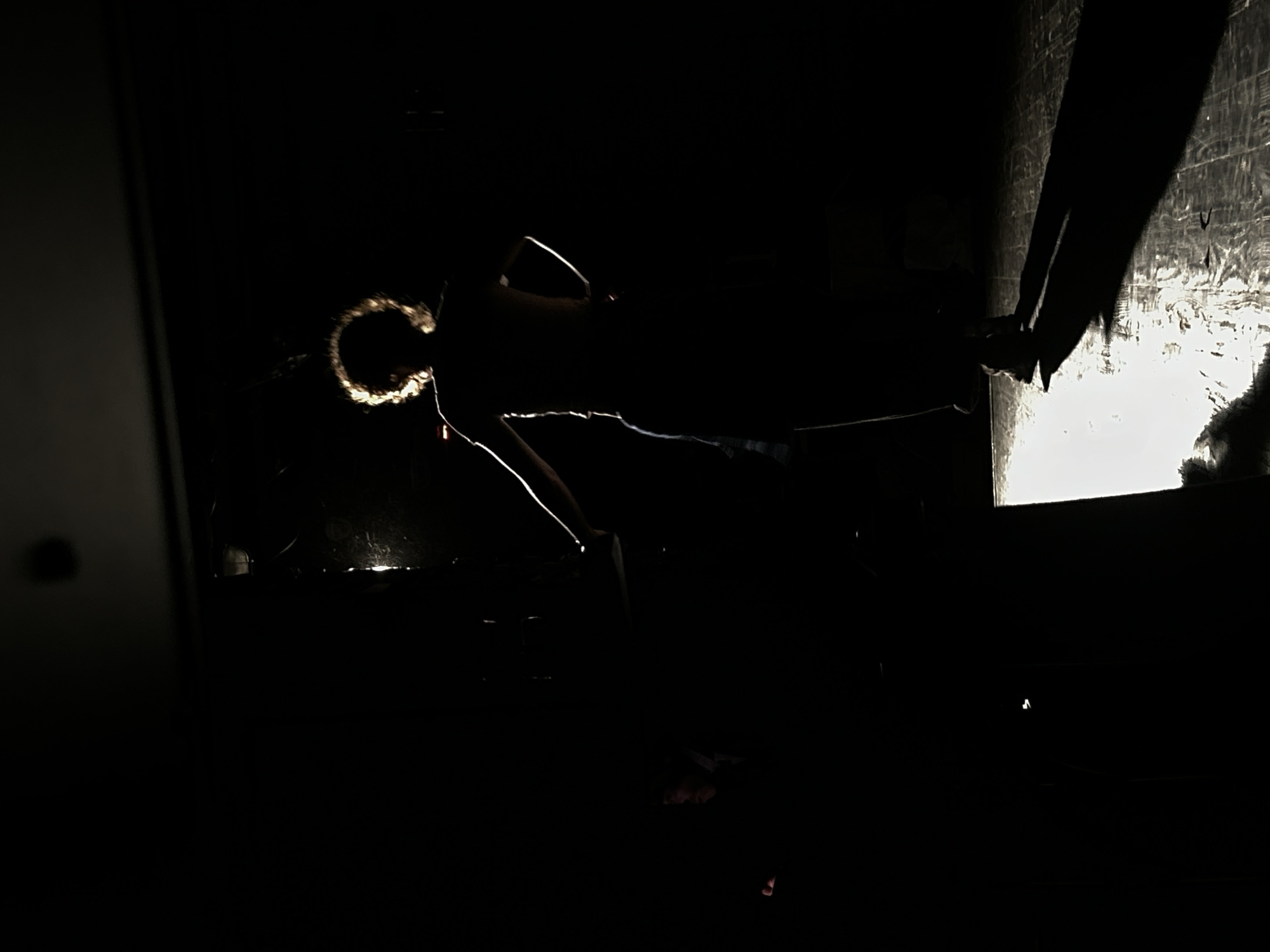 A silhouette of a person stands with their back to the camera, facing a bright light source. The light creates a halo effect around the person's curly hair and illuminates the edges of their body, casting a long shadow on the floor. The person is wearing a t-shirt and pants. The environment appears to be a dark room or stage, with the floor reflecting the light. The scene is lit with a dramatic, high-contrast lighting setup.