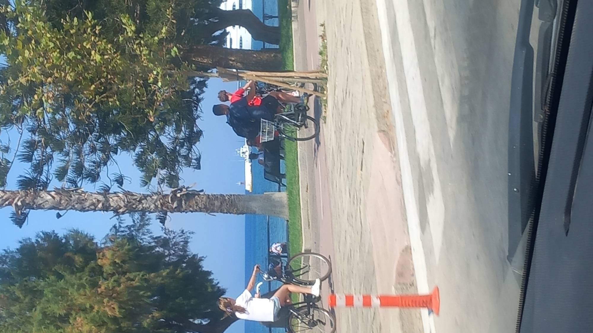 The image captures a scenic view of the city of Kos, Greece, likely along the waterfront promenade. Two individuals are seen cycling on a paved path that runs parallel to the sea. The person on the left is a woman with light-colored hair, wearing a white t-shirt and blue shorts, riding a black bicycle. The person on the right is a man wearing a dark t-shirt and black shorts, also on a bicycle, with a backpack. Behind them, the turquoise sea is visible, with a large white ship and a smaller yellow ship in the distance. The path is lined with trees, including a prominent palm tree, and some park benches are placed along the waterfront, with a few other people seated on them. An orange and white traffic cone is positioned on the left side of the path, in the foreground. The overall impression is one of a bright, sunny day with leisure activities taking place by the sea.