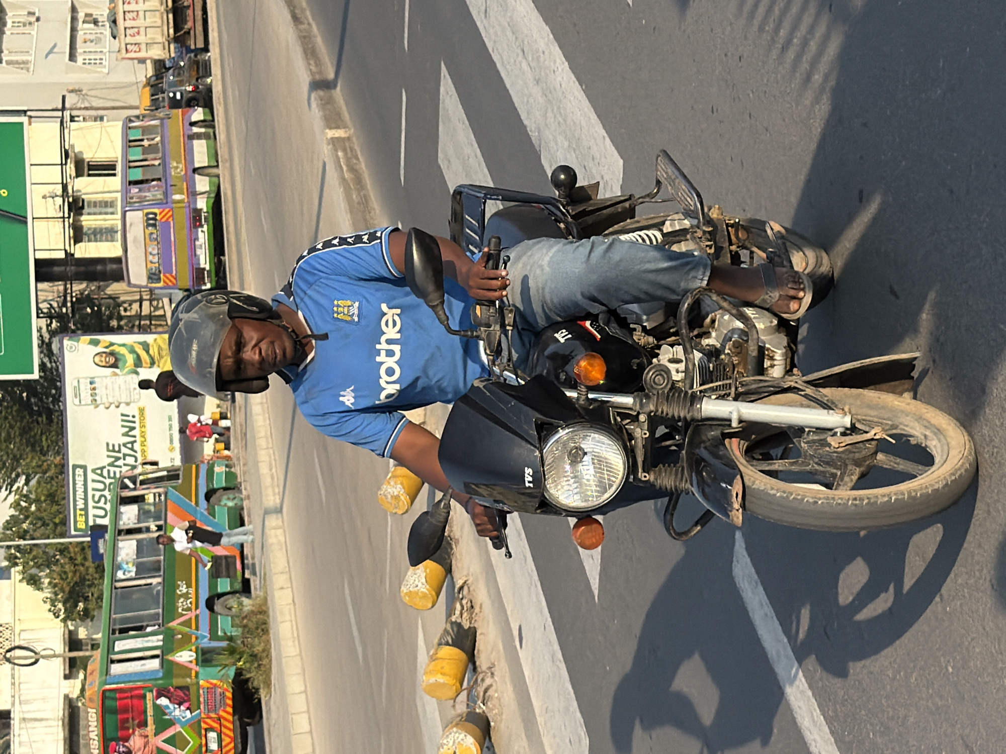 The photograph captures a street scene in Dar es Salaam, Tanzania, likely near a busy thoroughfare judging by the presence of public transport. In the foreground, a man wearing a blue shirt with the Kappa logo and the name "Brother" is riding a black motorcycle. He has a black helmet on and is looking forward. The motorcycle has a TVS logo on the front. The man is wearing blue jeans and brown sandals.
Behind the motorcycle, a brightly colored green and orange bus is visible, with passengers and advertisements on its side. To the right of this bus, another bus, purple and yellow, is also on the road. The background shows buildings and a large billboard with text, including "BET WINNER". Traffic cones, painted yellow and white, are scattered on the side of the road. The scene appears to be well-lit, suggesting it is daytime. The road surface is asphalt with white pedestrian crossing markings.