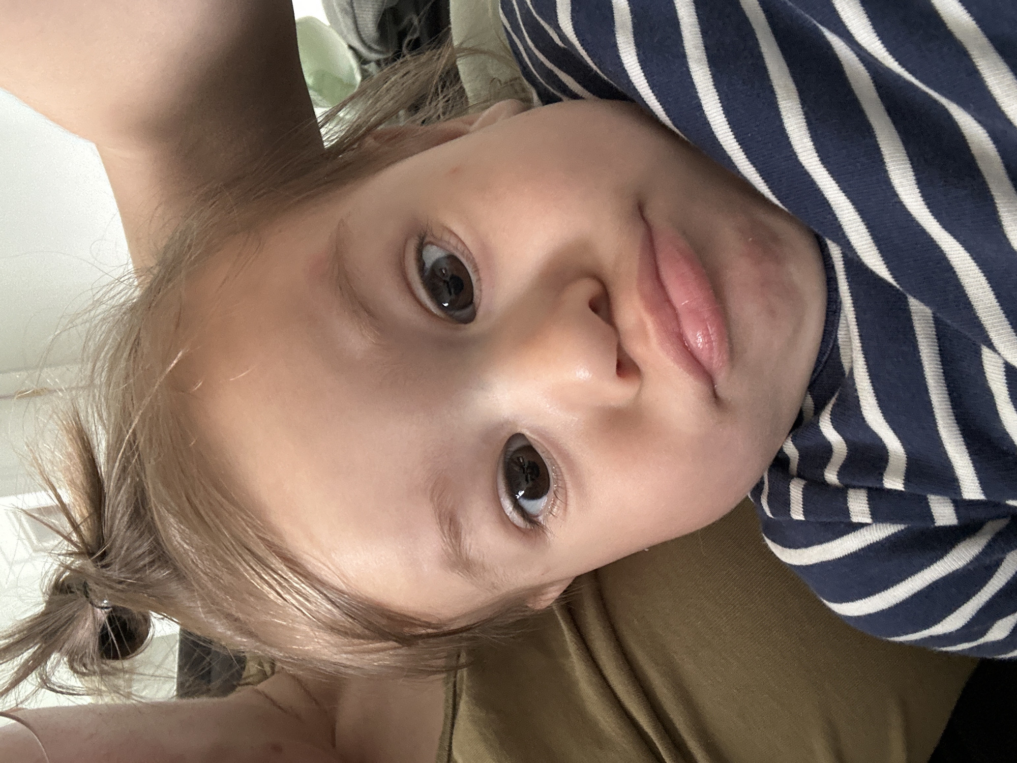 A young child with fair skin and light brown hair is lying down. The child is wearing a blue and white striped shirt. The child's hair is pulled back in a small ponytail on one side. The background appears to be blurry and includes a glimpse of green fabric. The photograph is taken at a close range. The child is looking directly at the camera. There is no indication of this picture being taken in Kharnavy, Russia.
