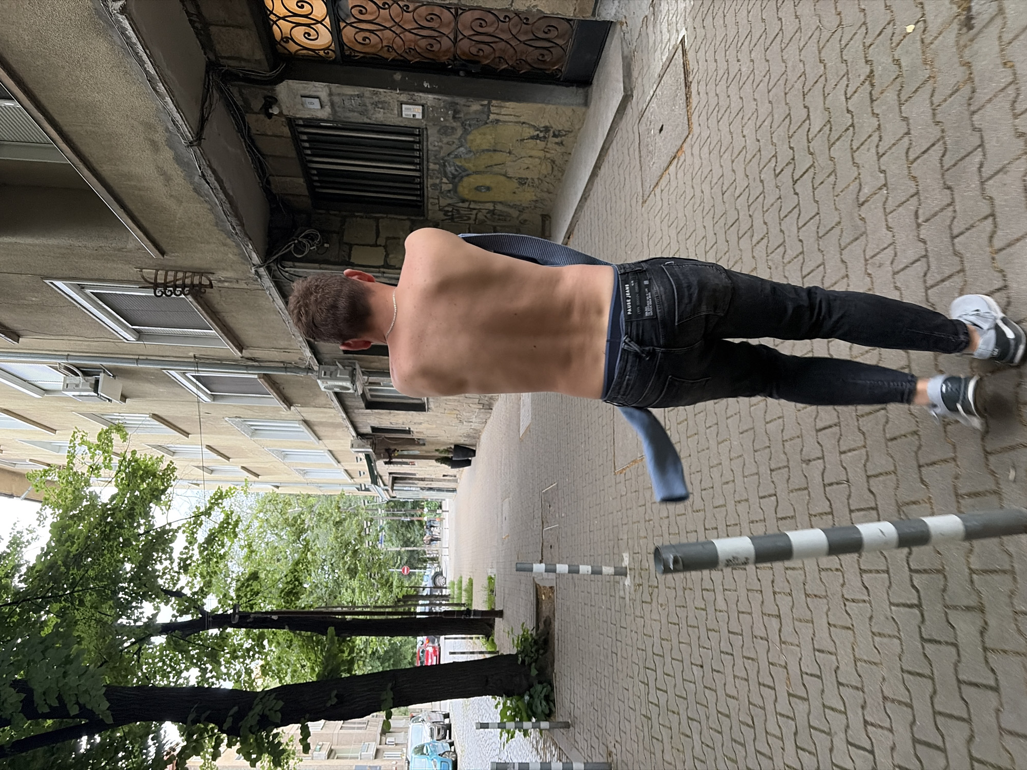 A person, from behind, walks down a paved sidewalk in an urban environment. The person is wearing dark jeans and grey sneakers, with their shirt pulled down around their waist. The sidewalk is made of interlocking grey rectangular pavers. To the left of the sidewalk are trees and the facade of a multi-story building with numerous windows. To the right of the sidewalk is the facade of another building, featuring a large, ornate dark metal door, graffiti on the lower section, and a prominent electrical conduit system running along the wall. The sky is overcast. In the background, more buildings and trees are visible, along with parked cars and other pedestrians.