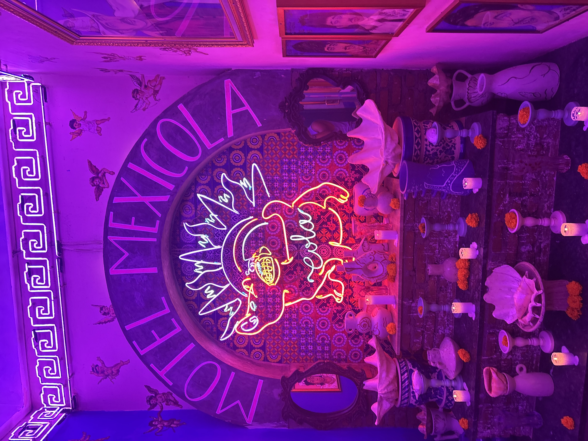 A darkly lit, vibrant scene is captured in what appears to be a themed interior space, illuminated by strong purple and pink neon lighting. The focal point is a large, arched neon sign that reads "MOTEL MEXICOLA" in bold, pink letters. Below this sign, a neon drawing of a sun-like figure with a horned head and a grinning mouth, eating spaghetti, is prominently displayed. The background behind the sign features a mosaic of colorful, intricate patterns.
On a tiered shelf in front of the sign, a collection of decorative objects is arranged. These include several white, shell-shaped bowls, various ceramic vases and vessels in shades of purple, white, and blue, and small, flickering white candles. Orange marigold flowers are scattered around the shelves, adding pops of color to the display.
To the left of the central display, a small, ornate, dark-framed mirror hangs on the wall, reflecting a glimpse of the interior. Above the neon sign, cherub-like figures are painted onto the wall, adding to the whimsical and slightly surreal atmosphere.
To the right of the main display, a series of framed portraits hang on the wall, also illuminated by the neon lights. The frames appear to be gilded.
The overall impression is of a stylized, artistic, and possibly festive environment, rich in color and decorative detail. The specific location in North Kuta, Indonesia, is not visually evident from the provided image itself, but the description of the space is consistent with a unique, themed establishment.