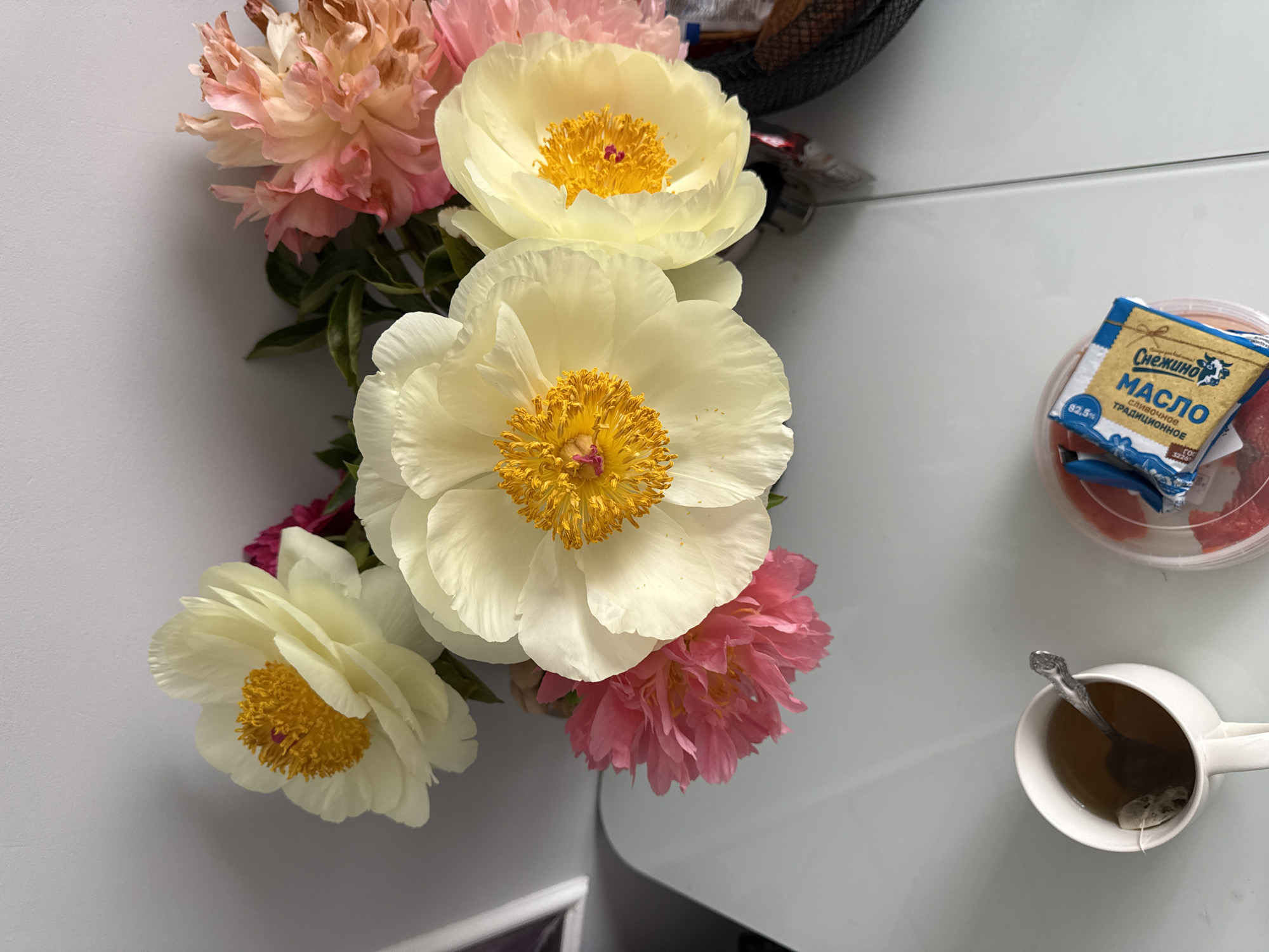 A high-angle shot shows a table with flowers, tea, and butter.
The table is white and reflective. Arranged on the table are some blooms of different colors: light pink, light yellow, and pink. Beside the flowers, there is a cup of tea with a spoon inside. A small package of butter sits atop a container filled with what appears to be sliced fruit. A basket is visible in the upper right corner.
The location and city are unknown.