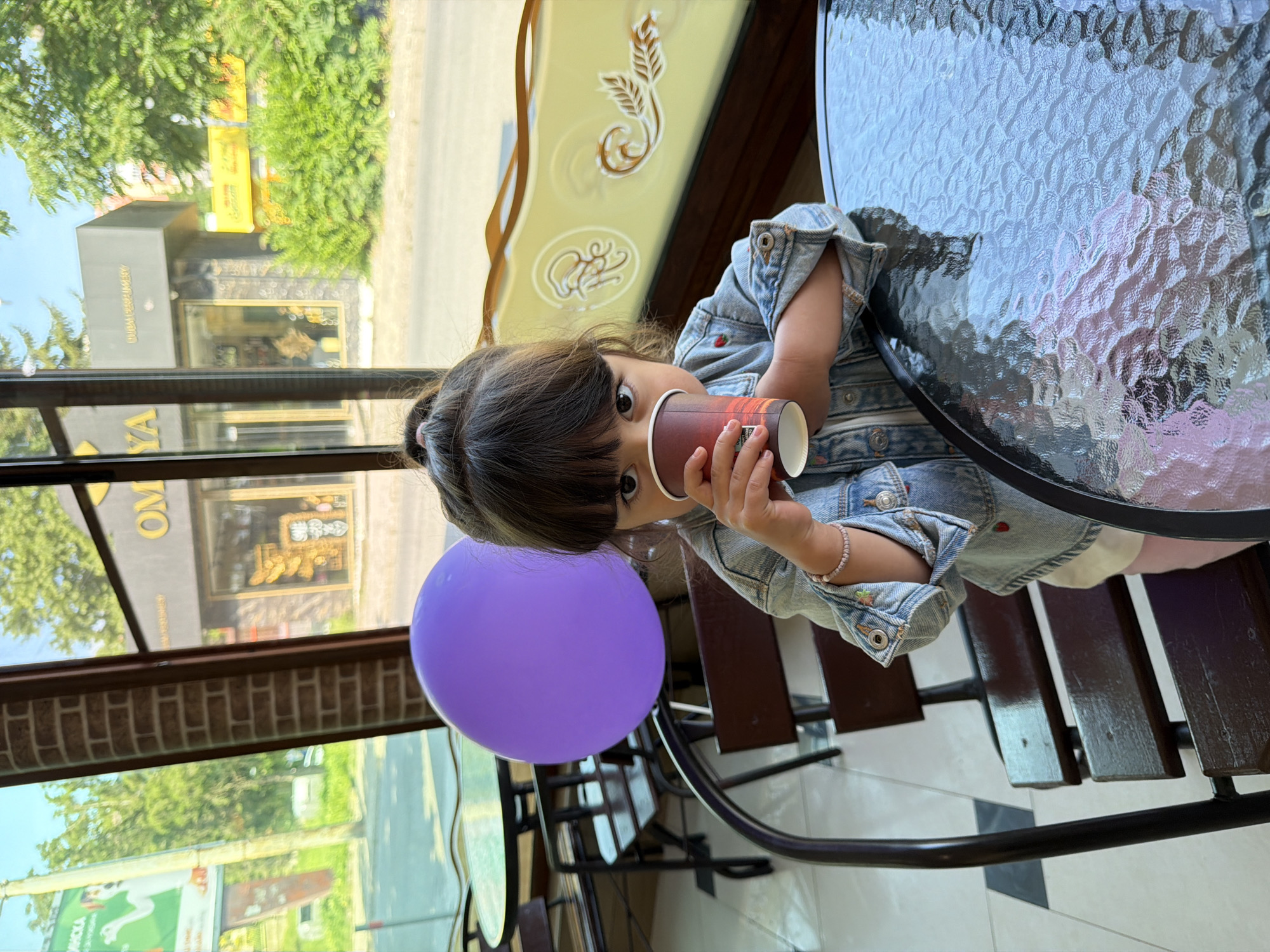 A young child, likely a girl, with dark hair tied in a top bun, is seated indoors at a cafe table, looking directly at the camera while drinking from a brown and red patterned paper cup held with both hands. She wears a light blue denim jacket with small colorful embroidered details (strawberries and flowers), a light-colored top, and light pink bottoms. A light pink beaded bracelet adorns her right wrist. A bright purple balloon floats behind her to the left. She sits on a dark wooden slatted chair at a round table with a textured, rippled glass top. The floor is tiled in a black and white checkered pattern. To her right, an ornate yellow panel with decorative gold patterns, including a stylized chef's hat or octopus figure and foliage, is visible.
Through the large window behind the child, a sunny urban street scene in Burgas, Bulgaria, is visible. Green trees line the street under a clear blue sky. Directly across the street, a dark-fronted building prominently features large gold Cyrillic letters spelling "ОМЯ" (OMYA), indicative of a cafe or shop, with display items visible in its window. To the left of this building, a partial green billboard with a cartoon dog figure and partially legible Cyrillic text (e.g., "НИККА") is seen. Further to the right, another dark building with a projecting sign displaying blurry gold text, and a yellow sign partially obscured by trees, completes the street view. The Cyrillic signage, particularly "ОМЯ," confirms the location as Burgas, Bulgaria.