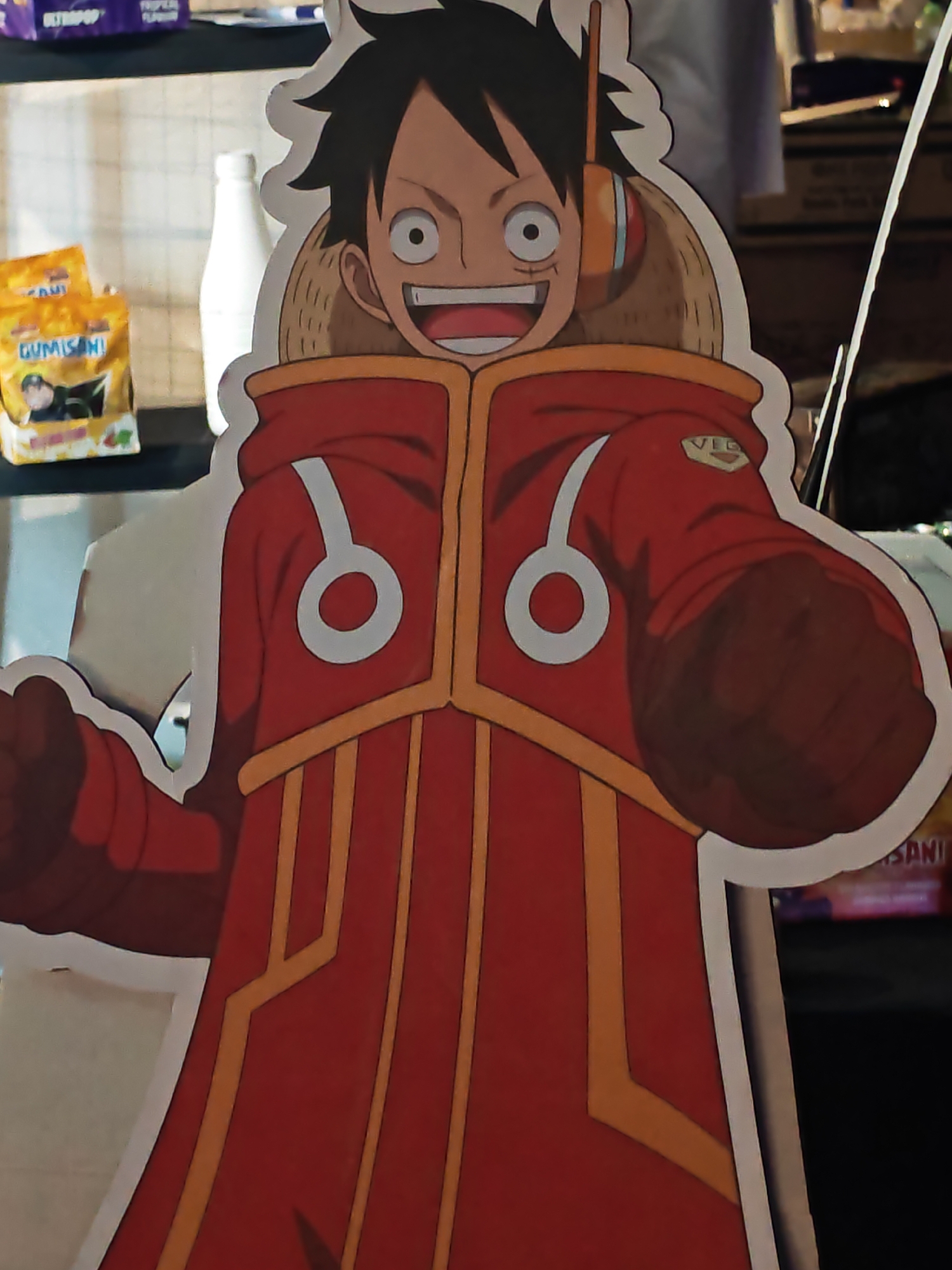 An anime character cutout, identified as Monkey D. Luffy from One Piece, stands in a display. The character is depicted wearing a red coat with yellow trim and white circular emblems. The cutout is positioned on a shelf, with various products visible in the background. To the left of the cutout are two yellow packages labeled "GUMISAN!". Behind the cutout, a white bottle is visible. The scene appears to be indoors, possibly a store or a collection display. The overall lighting suggests an artificial light source. The location is not specified in the image or provided context.