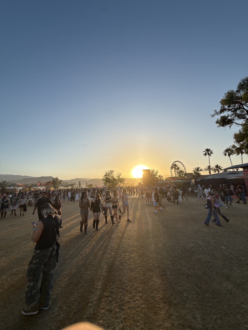 The image depicts an outdoor event, likely a music festival, taking place in Indio, United States, during sunset. A large crowd of people is visible, walking and interacting with each other on a mostly flat, grassy terrain. In the background, there's a Ferris wheel, stage structures, palm trees, and mountains silhouetted against the setting sun. The sky is clear with a gradient of blue to orange near the horizon. In the foreground, a person wearing camouflage pants and a vest stands with their back to the camera, holding a phone. The long shadows cast by the setting sun stretch across the ground, obscuring details of the landscape.