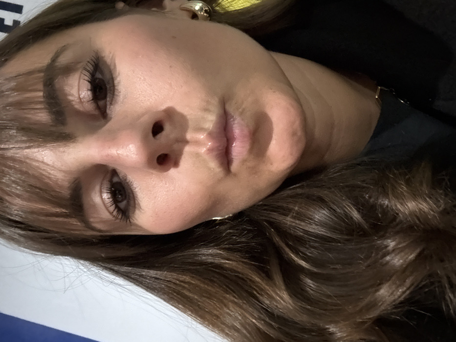 A close-up, angled portrait features a woman with her head tilted, looking towards the viewer. She has long, wavy brown hair with lighter highlights, styled with bangs that frame her forehead. Her dark eyes are open, with visible mascara on her lashes, and she maintains a neutral, calm expression. Her lips are closed, appearing glossy and a natural light pink. Her skin is clear, with a fair to medium tone.
She is wearing a chunky, gold-toned hoop earring in her visible ear, and a thin gold-toned chain or necklace is partially visible around her neck. The background is divided into a dark, shadowed area on one side, likely dark clothing or a backdrop, and a lighter white or light blue surface on the other, against which her head rests.
The woman is posing for the photograph. The lighting is soft and even, without strong shadows, making the time of day or specific weather conditions undeterminable. While the image does not provide explicit geographical markers, the context given is L'Étrat, France. Faint, indistinct blue text, possibly beginning with "L'E", is partially visible on the lighter background surface, obscured by her hair.
