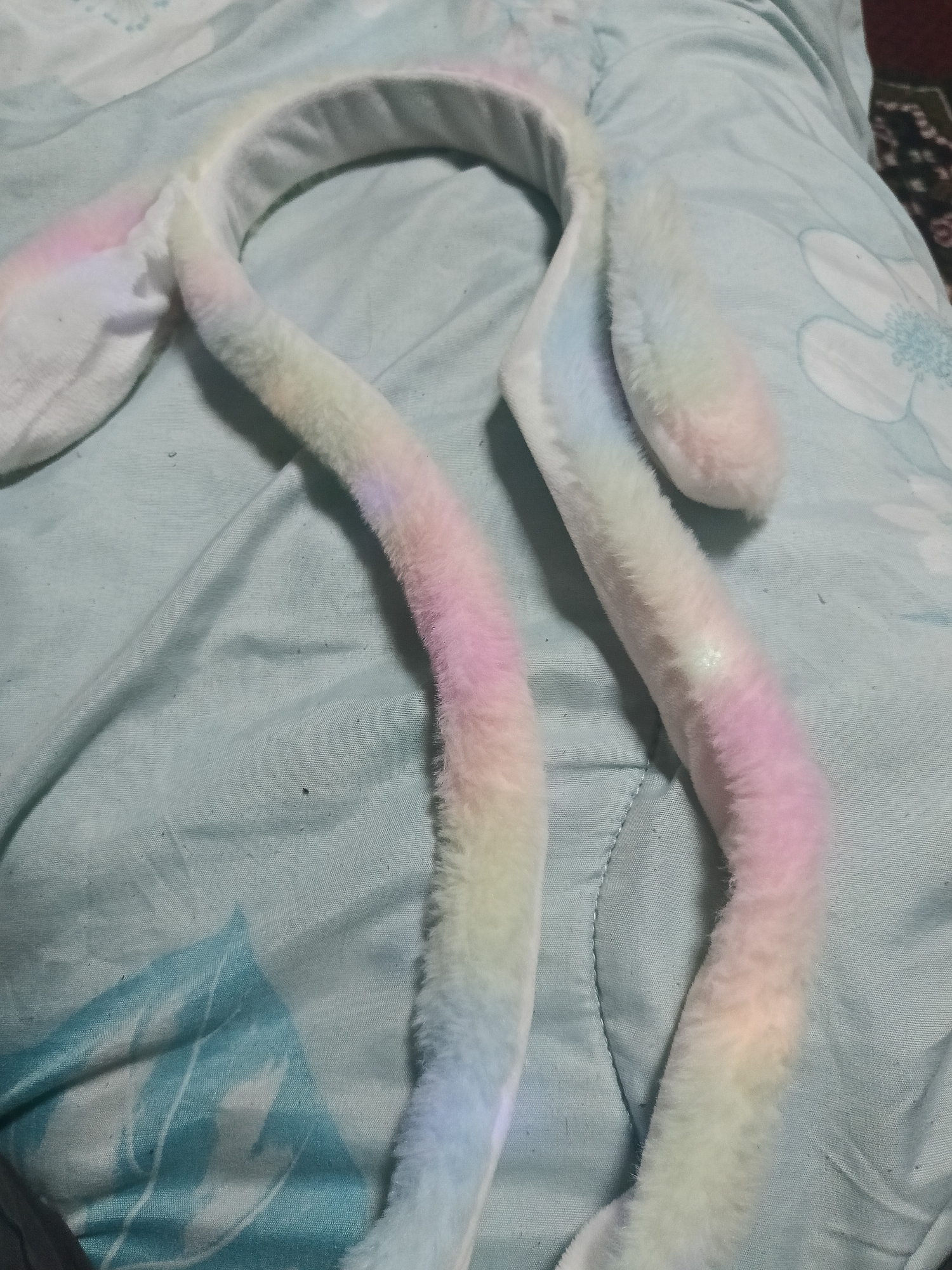 A fluffy, pastel-colored headband with rabbit ears is laid out on a light blue, textured surface, likely a blanket or bedspread. The headband is positioned diagonally across the frame, with the ears hanging down towards the bottom. The ears are soft and plush, featuring a gradient of colors including white, pink, yellow, and light blue. The headband itself is white and also displays subtle color variations. The surface beneath it has a stitched pattern, possibly of stylized flowers or leaves in a slightly darker shade of blue. The lighting appears to be ambient and diffused, casting soft shadows. No people or other animals are visible in the image. The scene is a close-up, focused on the headband. The location is not specified in Mizia, Bulgaria.