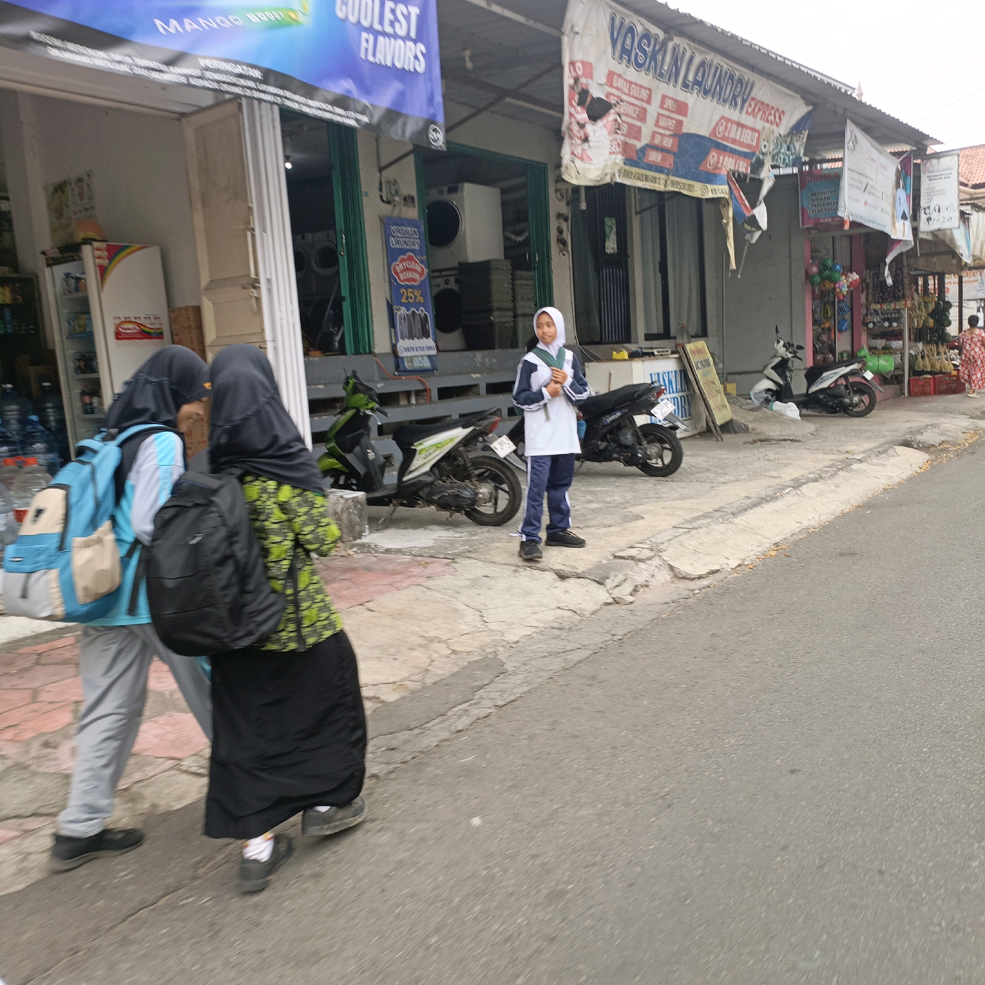 The image depicts a street scene in Salatiga, Indonesia, likely in a commercial area with storefronts. In the foreground, two individuals wearing hijabs are walking away from the camera. The person on the left is wearing a blue backpack and grey pants, while the person on the right is wearing a green patterned top and a black skirt, carrying a black backpack. To their right, another person in a white and blue hijab uniform stands, appearing to hold a book.
Behind the individuals, several shops are visible. One storefront has a large blue banner that reads "MANGO BOOST COOLER FLAVORS" and another banner that says "VASKLIN LAUNDRY EXPRESS." Another shop displays a sign for "VASKLIN LAUNDRY" with "DRY CLEAN" and "25%" visible. There are several motorcycles parked along the sidewalk, including a green and white scooter and a black motorcycle. Various items are displayed outside the shops, such as bottles of water, boxes, and some decorative items. The ground is paved with concrete and asphalt.
