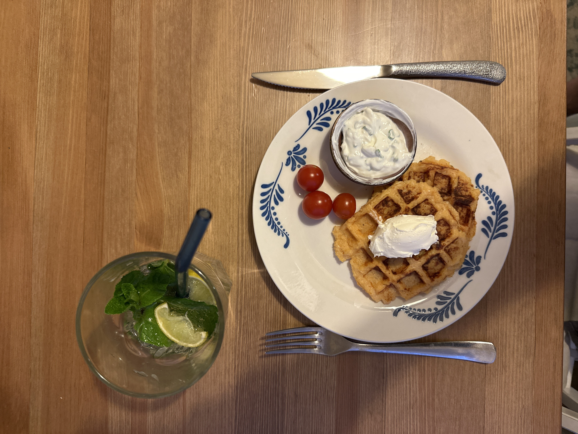 A top-down shot shows a wooden table set with a plate of food and a drink. The plate features two waffles, each topped with a dollop of white cream, a small bowl of a white creamy sauce with green specks, and three red cherry tomatoes. A knife is placed above the plate, and a fork is placed below it. To the left, a glass filled with a clear liquid, lime slices, and mint leaves sits with a blue straw. The location of the scene is not specified.