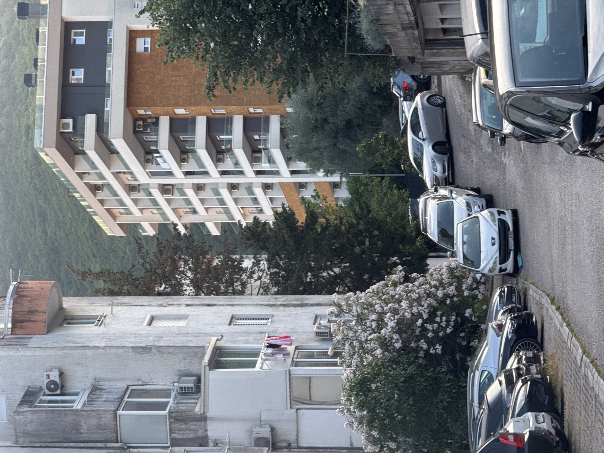 The image depicts a street scene in Budva, Montenegro. In the foreground, several cars are parked along a paved road. The cars vary in color and model, with some in the foreground being dark-colored and others further down the street being silver and white. To the left of the road, a tall, weathered apartment building with multiple windows and balconies dominates the scene. Laundry is hanging from a line attached to one of the balconies. Behind the apartment building, a modern, multi-story residential building with white facades and dark balconies stands tall. Further in the background, lush green trees and a mountain slope are visible under a hazy sky. The overall impression is of a residential area with a mix of older and newer architecture.