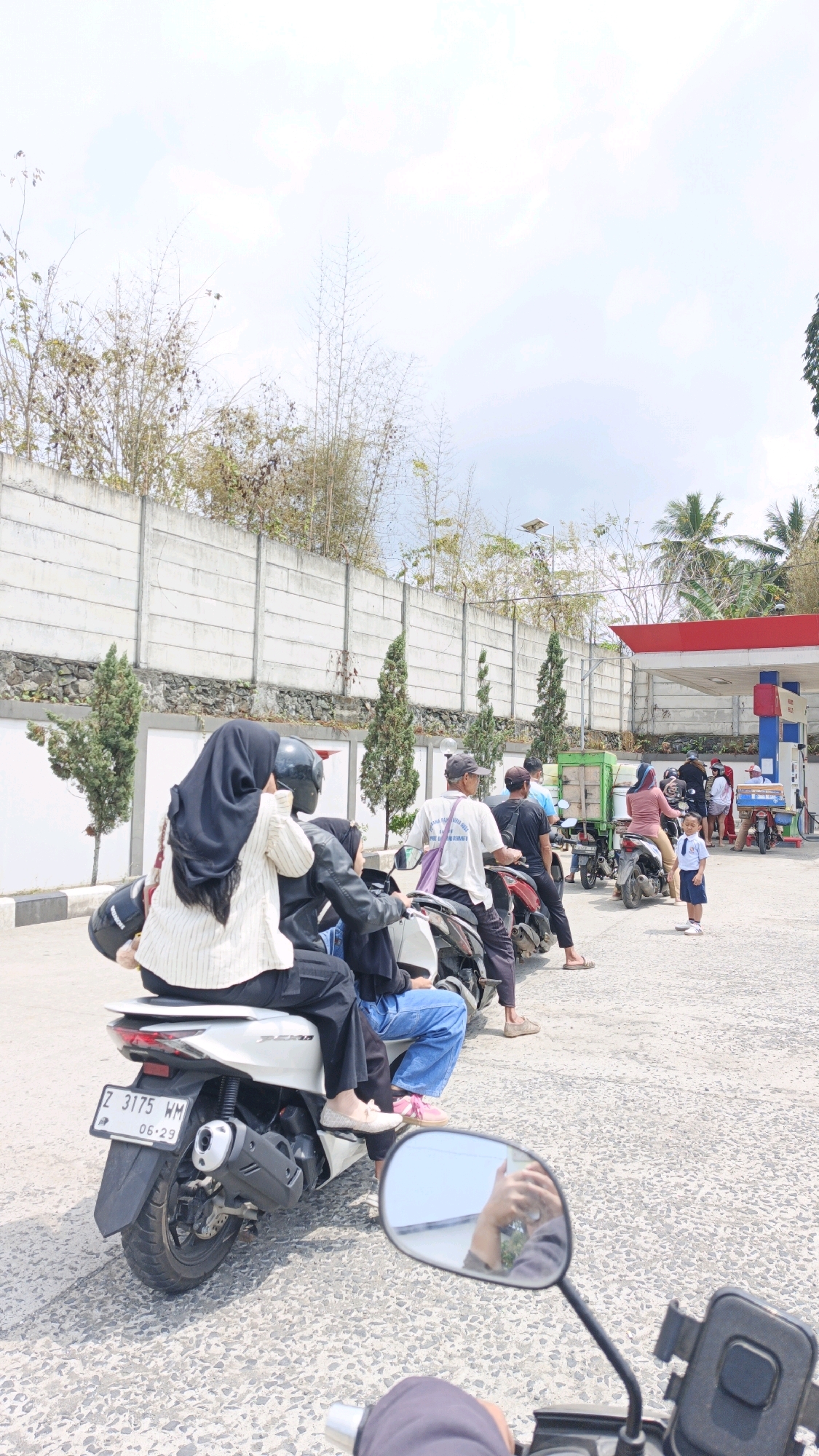 At a gas station in Sukaresik, Indonesia, a line of motorcycles and people are waiting to refuel. The scene is captured from the perspective of someone on a motorcycle, with the rear of their vehicle visible in the foreground. In the mid-ground, two individuals are seated on a white scooter, with the person in the back wearing a black hijab and a striped top, and the person in the front wearing a black leather jacket and blue jeans. Other people and motorcycles are in line behind them. In the background, a gas station canopy with red trim is visible, along with a white concrete wall and sparse trees. The ground is paved with asphalt. The sky is bright and hazy with some clouds.