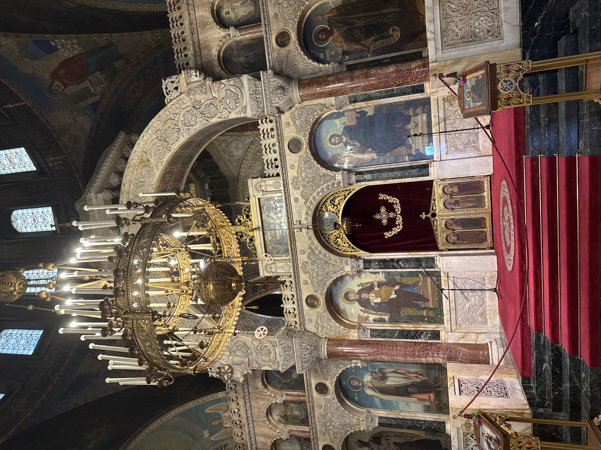 The image displays the ornate interior of the Alexander Nevsky Cathedral in Sofia, Bulgaria, characterized by its elaborate Orthodox Christian design. Dominating the upper center is a colossal, multi-tiered golden chandelier, adorned with numerous electric candles, illuminating the space. Beneath it, the grand iconostasis or altar screen forms the focal point, constructed from various stone textures and rich, carved ornamentation featuring intricate floral and geometric patterns.
The iconostasis is segmented by large, polished red marble columns and white carved pilasters, framing numerous arched niches that house large religious icons. Visible icons include a standing saint (possibly St. George or a warrior saint) on the left, the Theotokos (Virgin Mary) holding the Christ Child in the center-left, Christ Pantocrator holding an open book in the center-right, and Saint John the Baptist on the far right. Above these large icons, smaller arched windows and circular frames contain additional religious depictions. The upper sections of the walls and vaulted ceilings feature intricate frescoes and murals, including an angel in the upper-left corner and a draped figure in red on the upper-right arch. Arched windows with patterned grilles are visible high on the dark, vaulted ceilings, providing ambient light.
In the foreground, a set of red carpeted steps leads up to an elevated platform, also covered in red carpet. The stairs are flanked by dark, polished marble flooring. On the platform, a tall, ornate wooden icon stand with a golden base holds a framed icon depicting a saint, accompanied by a single red rose. To the left of this, a braided red rope acts as a barrier. A black microphone stand is positioned on the carpeted platform, and a small, gold-trimmed wooden screen with multiple icons is visible further to the left, framed by deep red velvet curtains that conceal the Royal Doors leading to the sanctuary. The entire scene is richly detailed with religious iconography and architectural craftsmanship, presenting a solemn and grand environment.