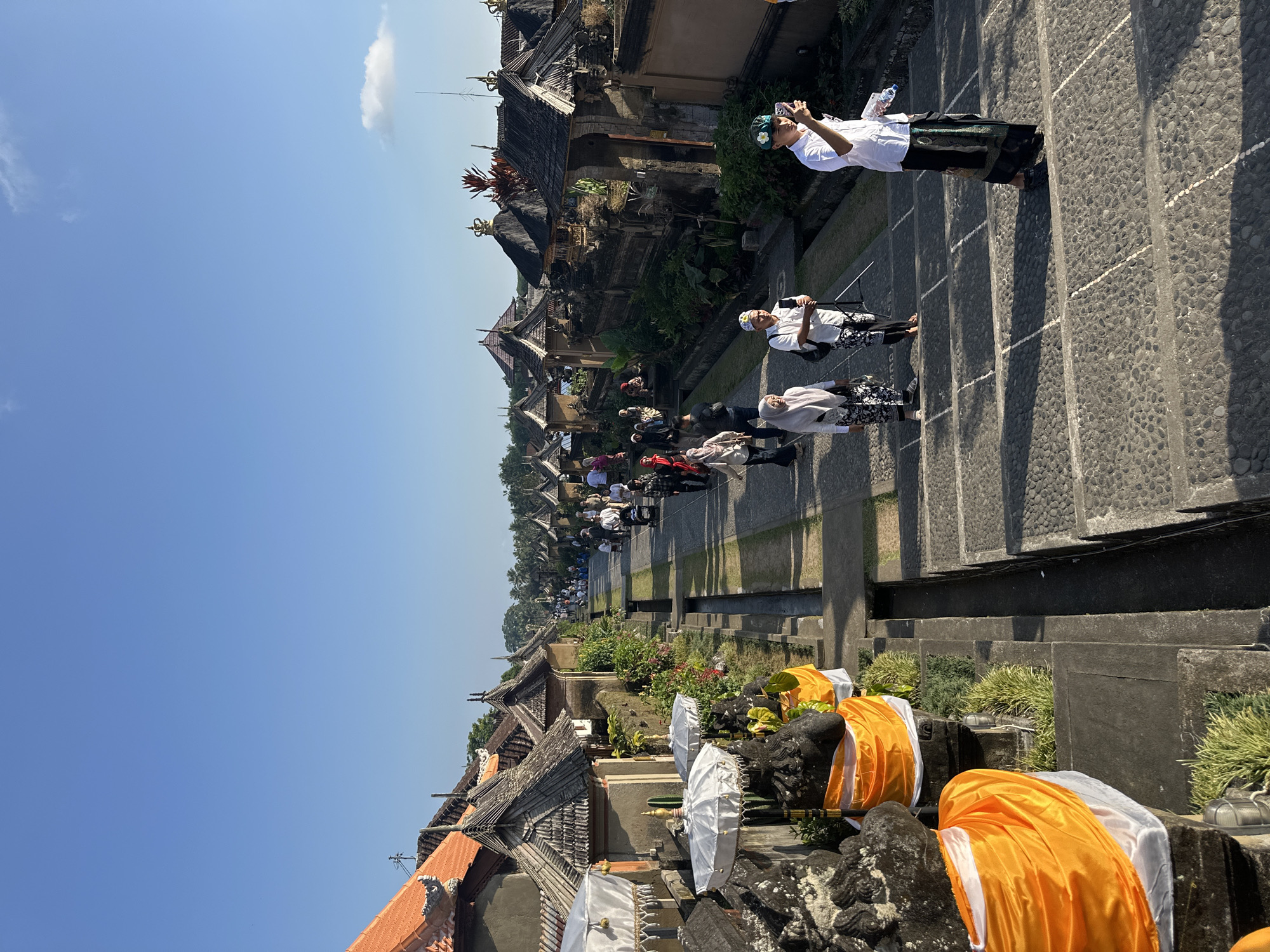 This image captures a sunlit scene along a stone pathway lined with traditional Balinese buildings in Bangli, Indonesia. The architecture features tiled roofs with intricate carvings and ornate golden finials. Several people are walking along the pathway, some dressed in traditional attire, including sarongs and head coverings. One individual in the foreground on the right is holding up a mobile phone, appearing to take a photograph. White umbrellas adorned with fringe are positioned on stone structures to the left, draped with bright orange fabric. Lush greenery and tropical plants are visible throughout the scene. The sky is a clear, bright blue with a few wispy clouds. The pathway appears to be a set of stairs made of dark cobblestones, with lighter lines marking the edges of the steps. A narrow channel, possibly for water drainage, runs alongside the pathway. The overall impression is of a cultural site or village, bathed in the warm light of day.