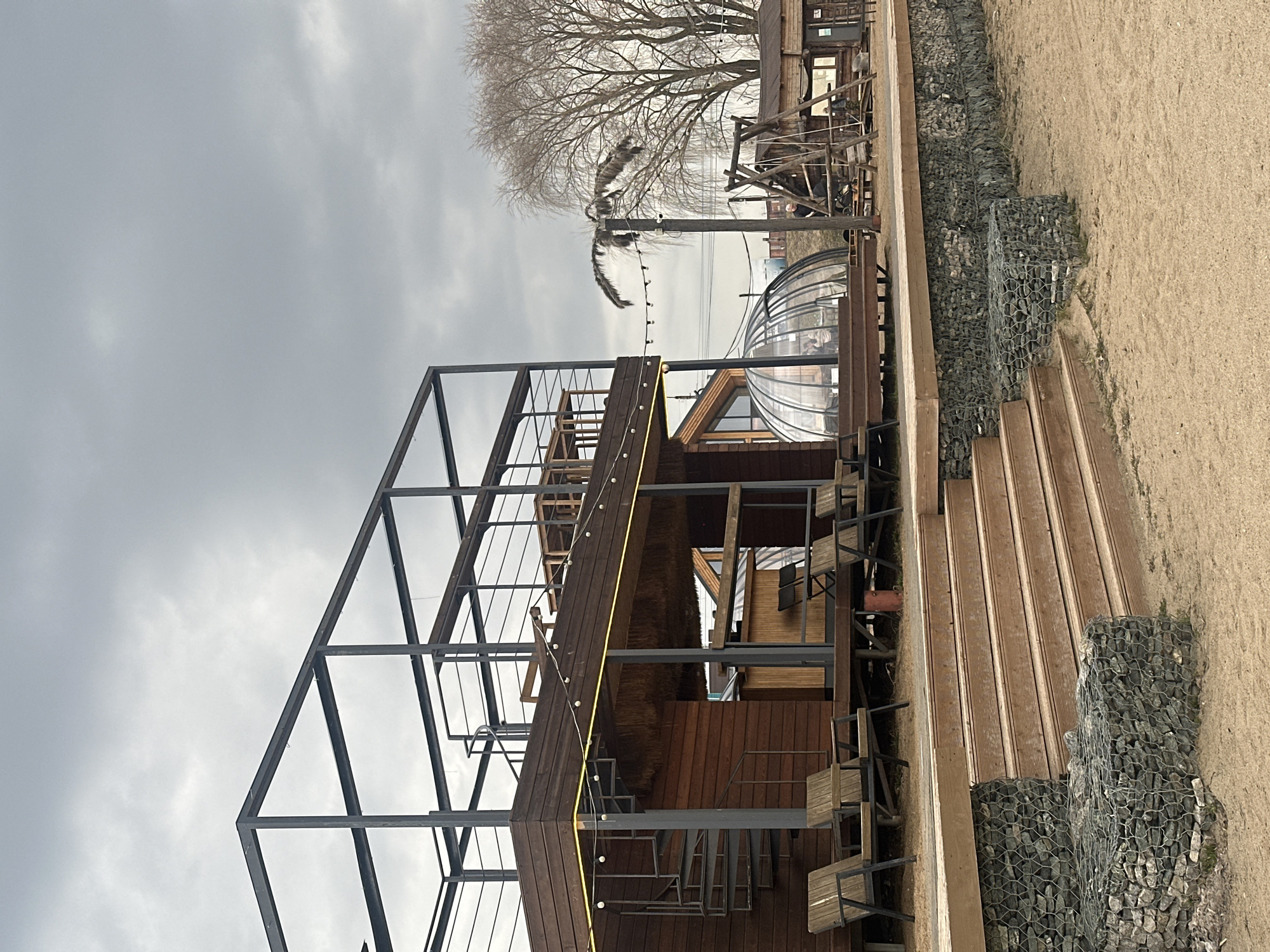 The image depicts a multi-level wooden structure with metal framework, possibly a restaurant or recreational facility, located outdoors near a sandy area or beach. The sky is overcast. There are wooden stairs leading up alongside gabion walls. A string of lights is visible along the structure. A geodesic dome-like structure is also present. Based on the presence of "Votkinsk" in the prompt, the location is likely Votkinsk, Russia. No people are clearly visible.
