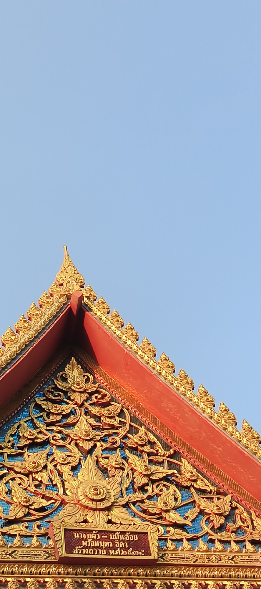The image displays a close-up, upward-angled view of the ornate roof of a traditional Thai temple or building against a clear, bright blue sky. The prominent features are the vibrant colors and intricate golden decorations, illuminated by natural daylight.
The roof is characterized by a steep, red-tiled gable, topped with a golden spired finial. Along the edges of the red roof, numerous smaller, highly decorative golden elements, resembling stylized flames or foliage, are arranged symmetrically.
In the triangular pediment section below the main roof, there is a large, elaborate bas-relief carving crafted in shimmering gold. This intricate artwork features traditional Thai motifs, including swirling patterns and stylized floral or mythical creature designs, set against a rich blue background.
At the lower center of this golden pediment, a small red plaque with white Thai script is visible. The text reads: "นาง แดง - แปรนล้อม พ่อแม่อุปถัมภ์ สร้างถวาย พ.ศ.๒๕๖๕". This translates to "Mrs. Daeng - Plaenlom, parents' supporter, donated/created in B.E. 2565," indicating a donation or construction date corresponding to the year 2022 AD. The architectural style and script confirm the setting in Thailand, specifically Bangkok as per the context. No people or activities are present, the focus is solely on the architectural details.
