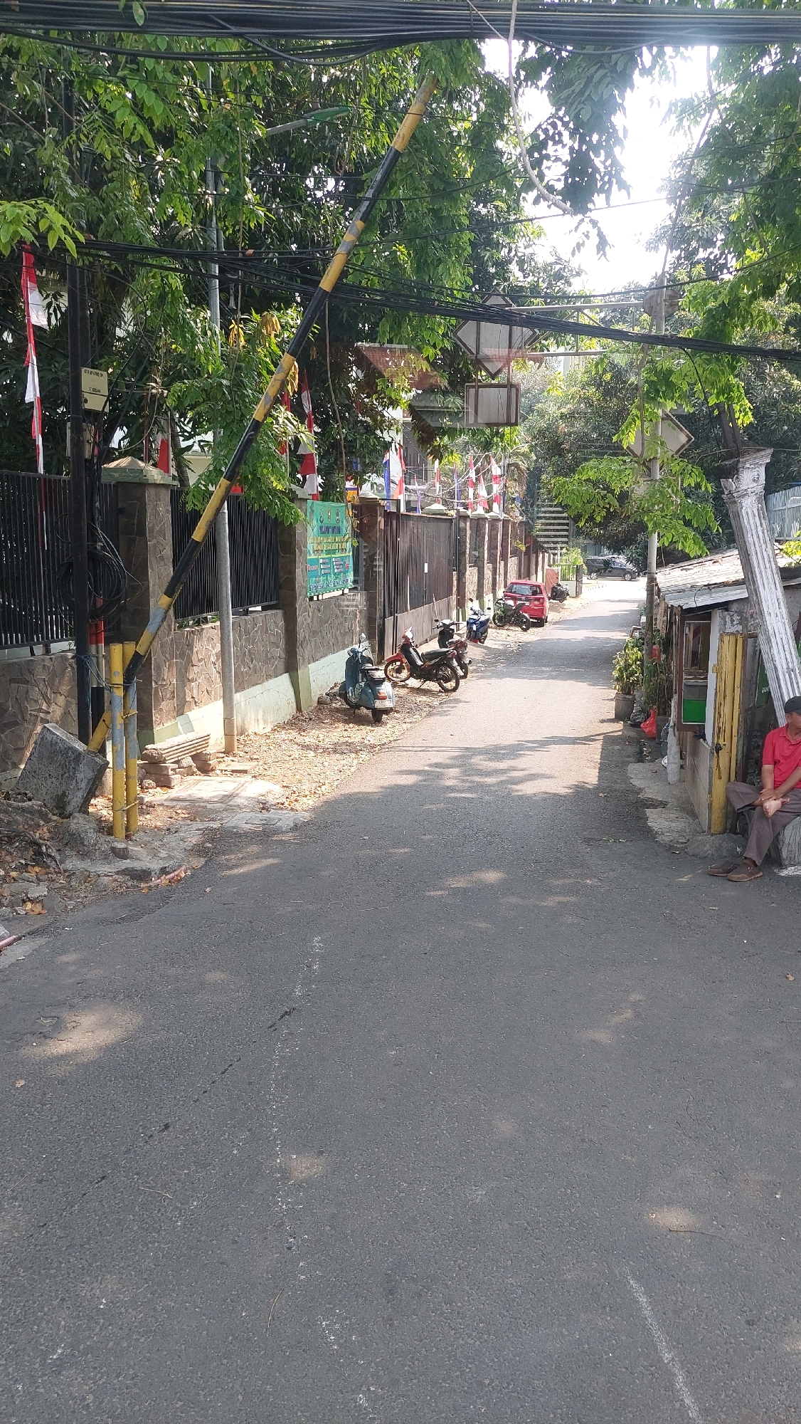 The image captures a street scene in East Jakarta, Indonesia, under a clear sky. A black and yellow striped boom gate is partially lowered across the road. The street is paved with asphalt and shows signs of wear, with some cracks and faded white markings. On the left side of the street, a stone wall borders a property with a black metal fence. Several Indonesian flags, red and white, are visible along the fence. A concrete barrier is placed on the sidewalk near the boom gate. Parked along the side of the road are several motorcycles, including a blue scooter and two darker motorcycles. A red car is parked further down the street. On the right side of the road, a small, open-fronted wooden structure with a white roof is situated. A man wearing a red shirt and dark pants is seated on a concrete ledge next to this structure. The scene is lined with trees, their leaves creating dappled shadows on the road. Debris, including dry leaves and small branches, are scattered on the ground along the edges of the street. Overhead, a dense network of electrical wires is strung between poles.