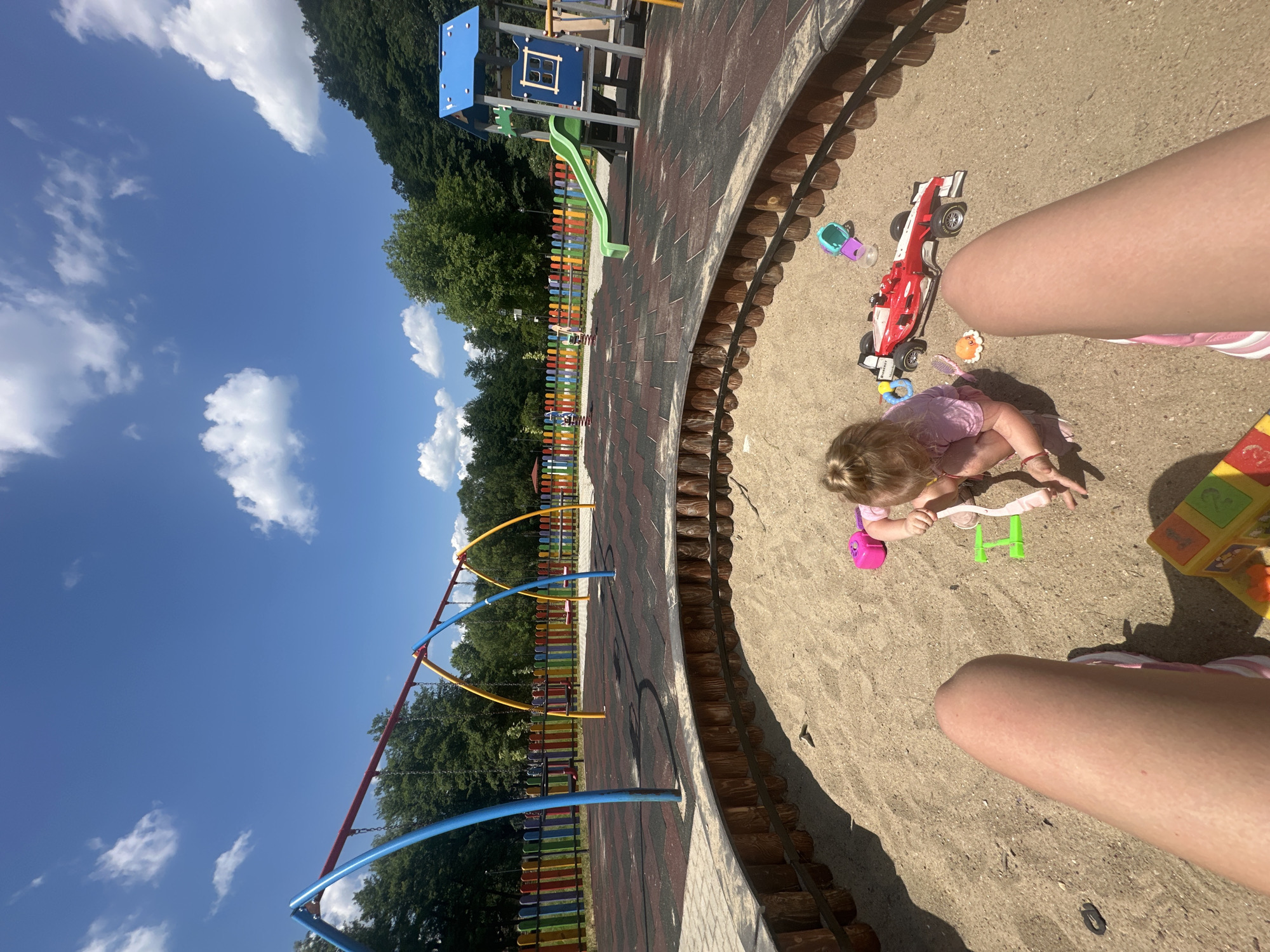 This is a medium shot of a child playing in a sandbox at a playground. The child is a toddler with blonde hair, wearing a pink shirt and shorts. They are surrounded by toys, including a red and white race car, a pink spade, and a green rake. The sandbox is circular and made of wood. In the background, there is a swing set, a slide, and a colorful fence. The playground is located in a park with trees and a blue sky with white clouds. The location is the playground in the city of Donchovci, Bulgaria.