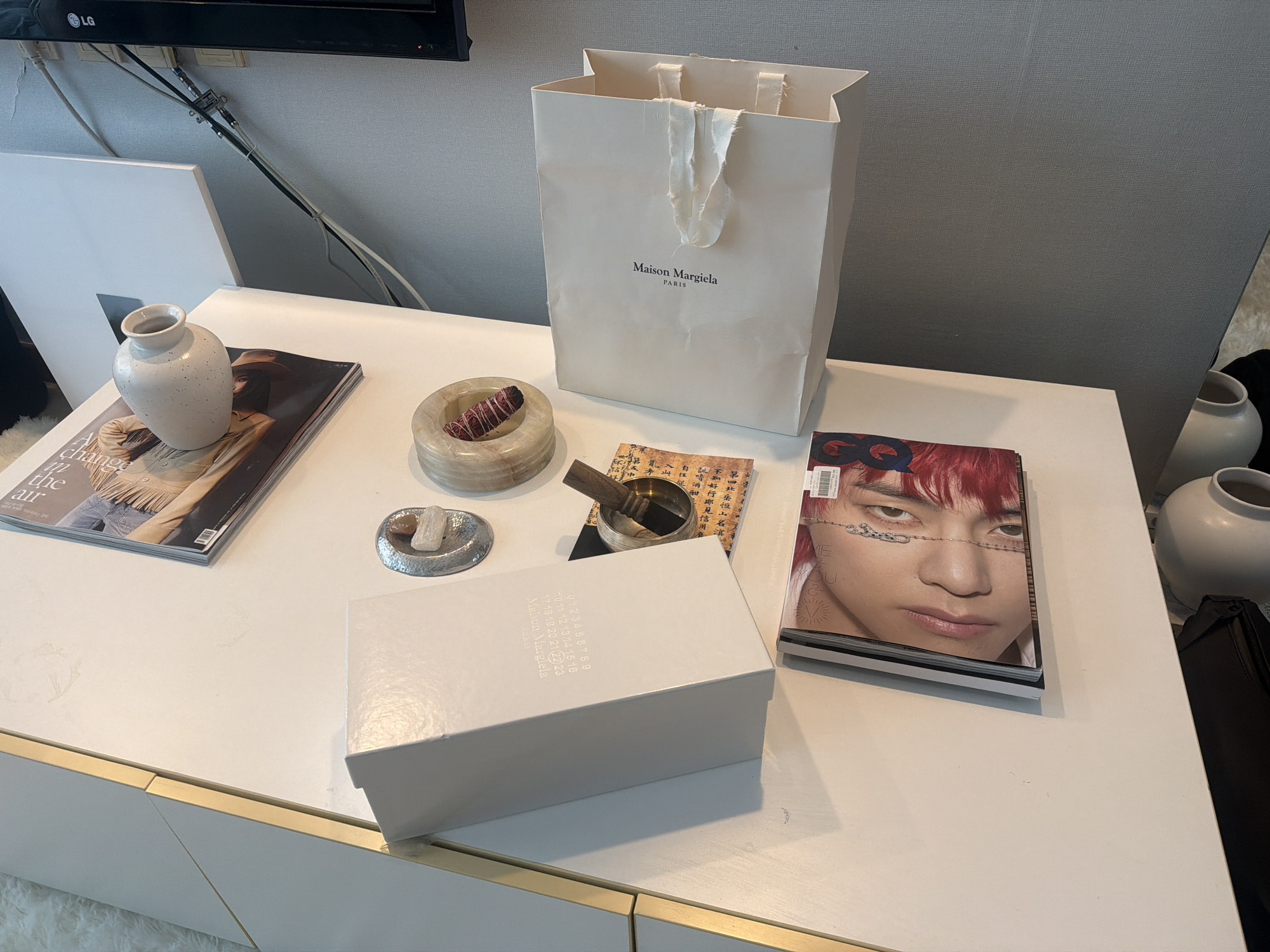 This image captures a curated display on a white, modern console table with gold accents in an indoor setting. The scene is bright, suggesting daytime lighting.
The central object is a white Maison Margiela shopping bag with fabric handles, bearing the "Maison Margiela PARIS" logo. Adjacent to it, a white rectangular box, also from Maison Margiela, displays a series of numbers from "0" to "23" on its top surface.
Various decorative items and magazines are arranged on the table. To the left, a speckled white ceramic vase rests on an open magazine featuring a person in a cowboy hat and the partial text "Change in the air." Nearby, a round, light-colored stone bowl contains a reddish sage bundle. A small metallic bowl with a wooden item (possibly a singing bowl or incense burner) sits on a piece of paper with visible East Asian characters. A small, textured silver dish holds a few small stones.
To the right, a stack of magazines is topped by one prominently featuring a person with red hair and distinct eye makeup on its cover, with the "GQ" logo visible. A barcode sticker is also present on this cover.
In the background, an "LG" branded television is mounted on the wall, with cables neatly arranged below it. A white canvas leans against the wall on the left, and another simple white vase is partially visible on the right, suggesting additional decor in the room. The overall impression is one of a clean, minimalist space with a focus on designer items and contemporary aesthetics, common in stylish residences in places like Seoul, South Korea, as contextually provided. No human activity is depicted; it's a static arrangement.