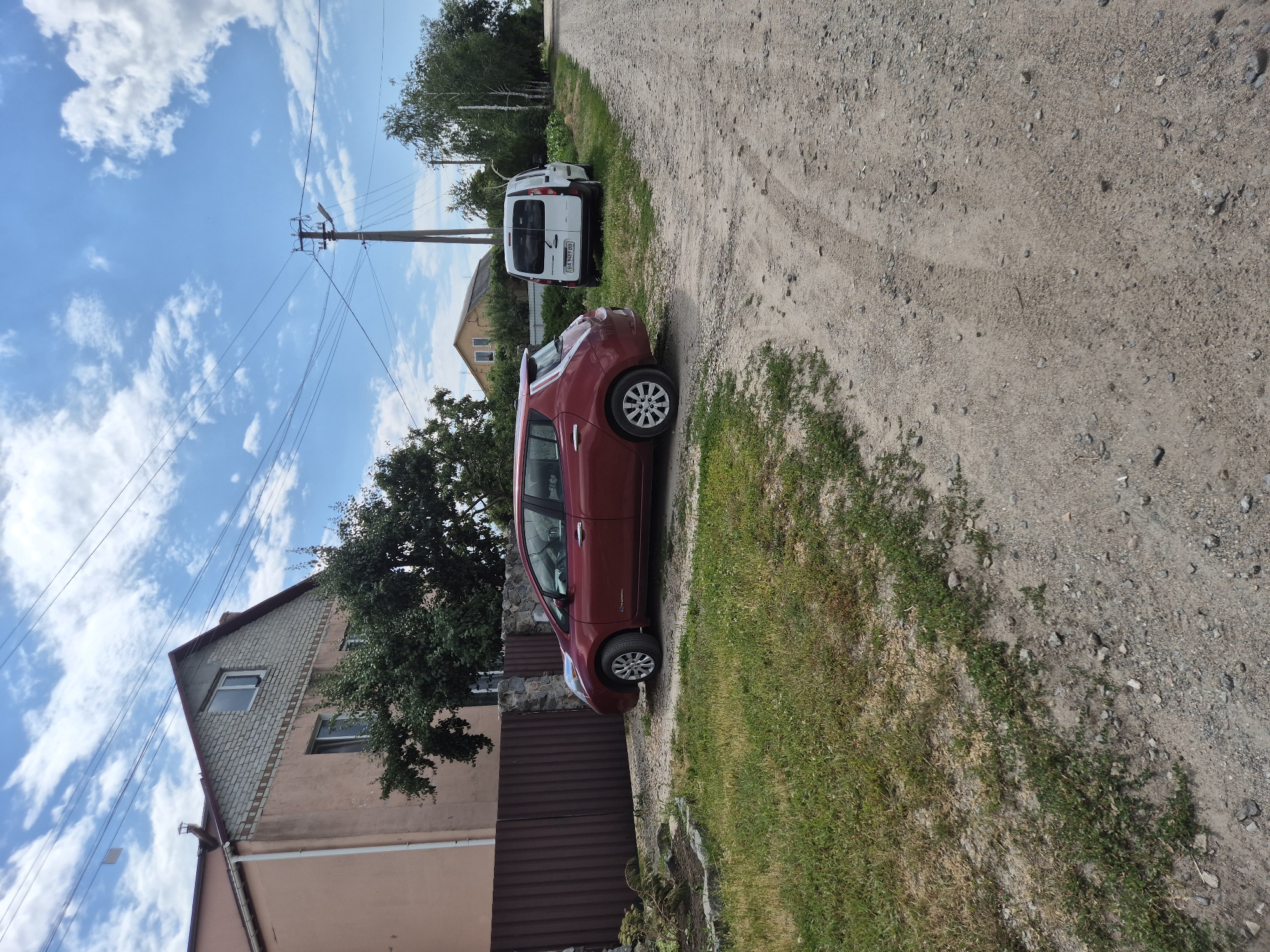 The image depicts a rural scene in Sozonivka, Ukraine. Two cars are parked on a dirt road next to a house and a grassy area. A red hatchback is parked in the foreground, while a white van is parked further down the road. The house is a multi-story building with a light pink facade and a brick upper section. There are power lines and a utility pole visible in the sky. The sky is blue with scattered white clouds, suggesting it is daytime. The overall impression is of a quiet, everyday scene in a village. The license plate on the white van is partially visible and appears to read "BI 949 BB".