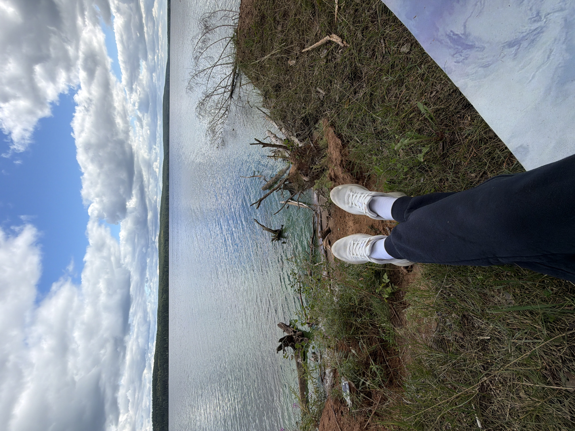 The image shows a first-person perspective of a person sitting on the grassy bank of a large body of water. The person is wearing white sneakers and dark pants. The water is calm with some ripples, and the sky is partly cloudy with blue patches. A line of trees borders the far side of the water. In the foreground, there are some fallen branches and vegetation on the bank. The location is identified as being in the city of Chaikovsky, Russia.