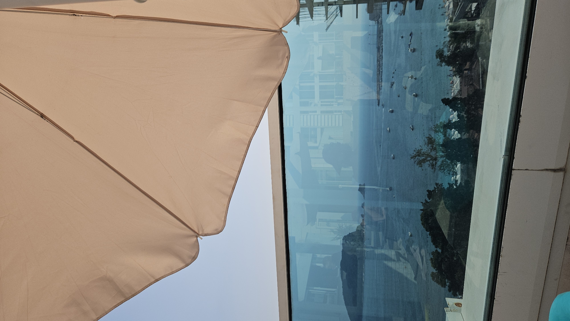 The image captures a view from a balcony or patio, looking out over a body of water. A large, light-colored umbrella is angled from the top right of the frame, providing shade. Below the umbrella, a glass railing separates the viewing area from the scene beyond. Through the glass, a calm, blue sea stretches out, dotted with several small boats. In the distance, a rocky island or peninsula rises from the water, with trees and buildings visible on its slopes. Reflections are visible in the glass, including a silhouette of a person wearing a cap. The lower portion of the image shows some foliage and buildings, suggesting a coastal town or resort. The sky is clear and pale blue.