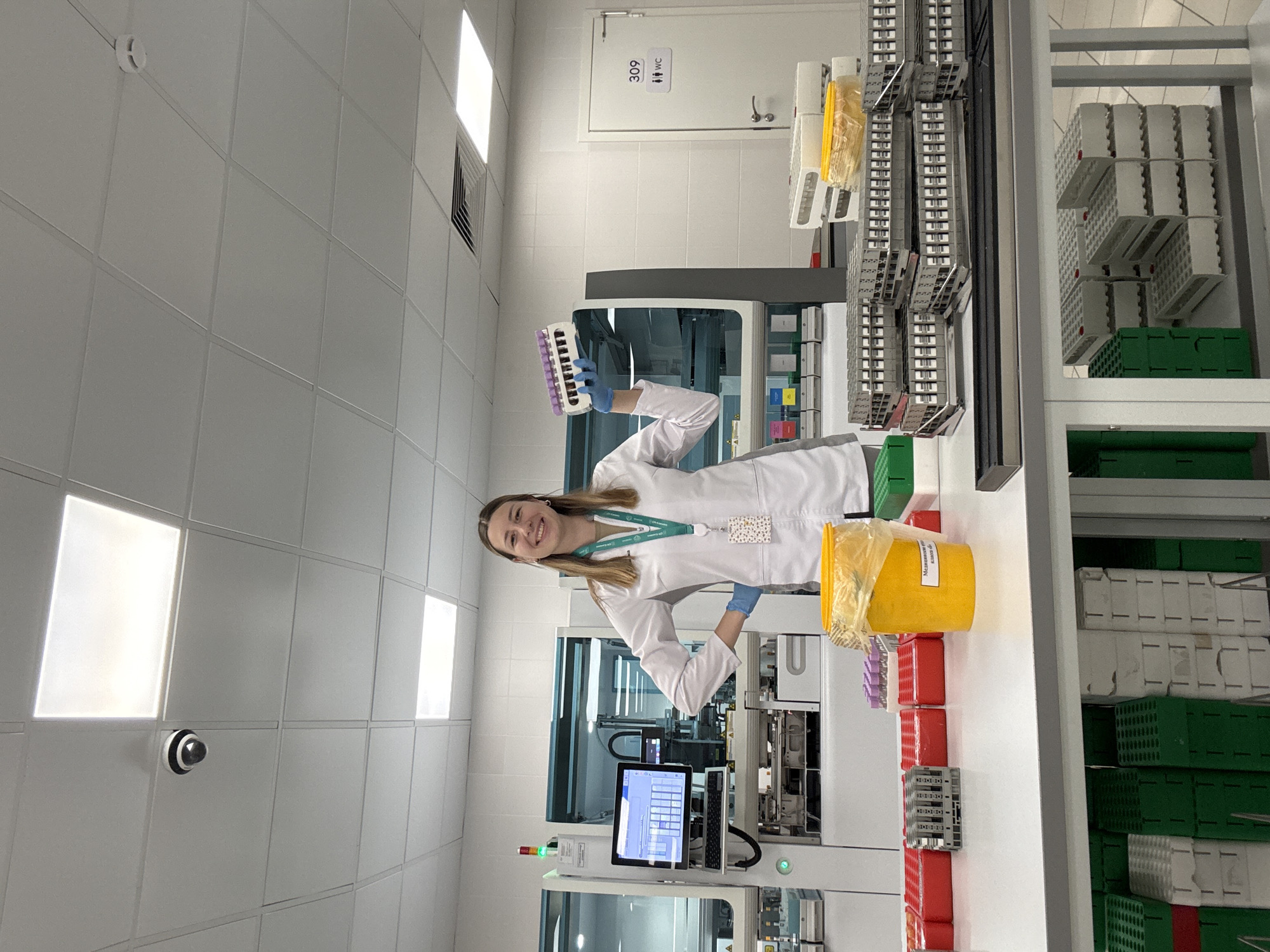 A young woman in a white lab coat and blue gloves stands in a laboratory, smiling at the camera. She holds a rack of test tubes in her right hand. To her left is a large piece of laboratory equipment with a computer screen displaying a graph. In front of her, on a table, are several racks of test tubes, a yellow bin, and other laboratory supplies. The room is brightly lit with fluorescent ceiling lights and has white tiled walls. A security camera is visible on the ceiling. A door with a sign indicating "309 WC" is visible in the background. The overall scene suggests a working laboratory environment.