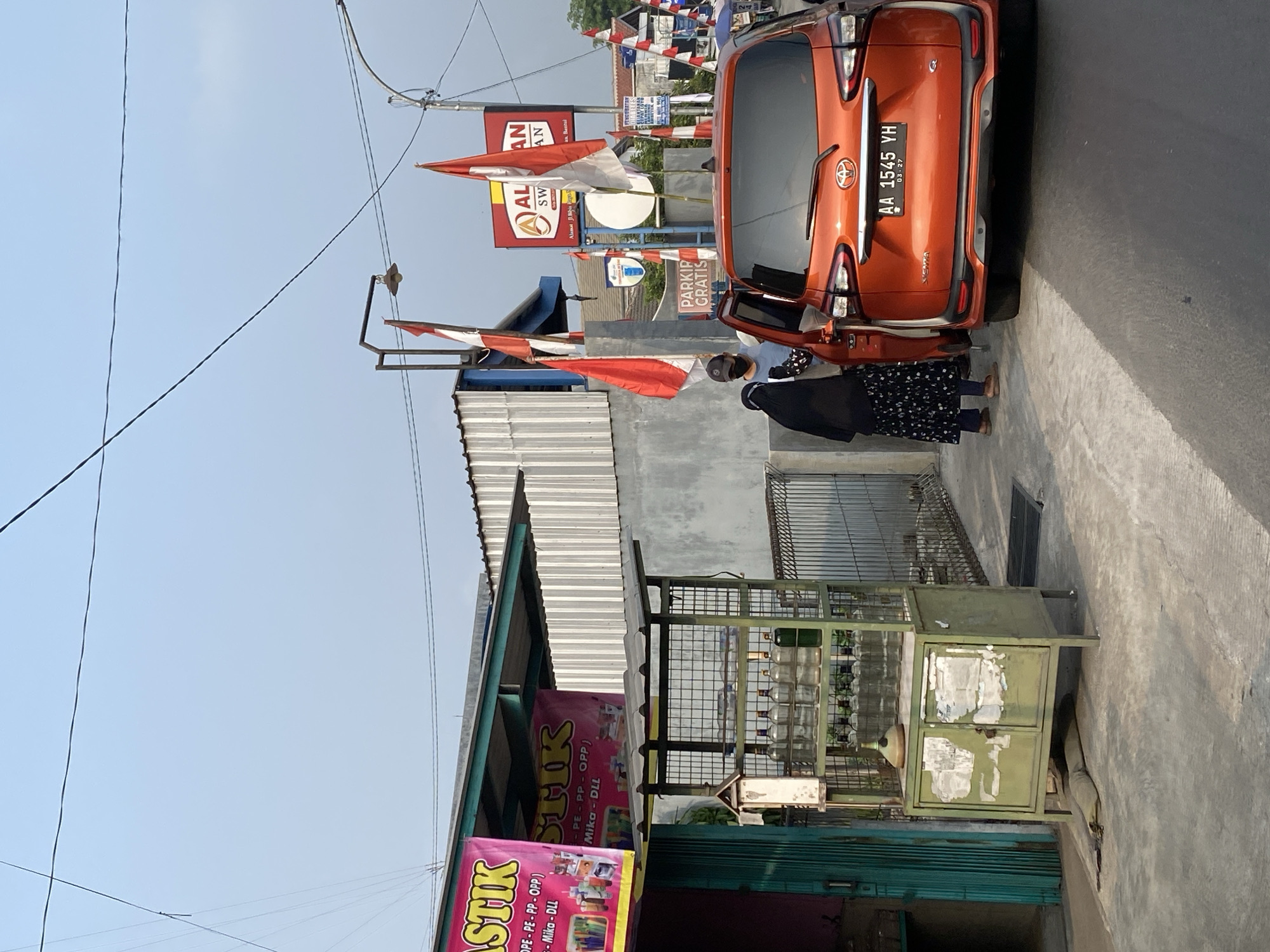 An orange Toyota Sienta is parked on the side of a road in Kasihan, Indonesia, with its rear right door open. A person wearing a black hijab and a patterned skirt stands at the open door. Another person, wearing a light blue t-shirt and a dark baseball cap, is leaning into the car from the driver's side. Two Indonesian flags are visible, one on a pole behind the car and another smaller one mounted on a nearby structure. A sign with text in Indonesian, reading "ALAN SWAN," is visible in the background. To the left, a green metal rack displays rows of glass bottles, possibly for sale. A pink banner with text in Indonesian advertising "PLASTIK" is attached to the awning of a building. The scene appears to be set in a commercial or roadside area under a clear sky.