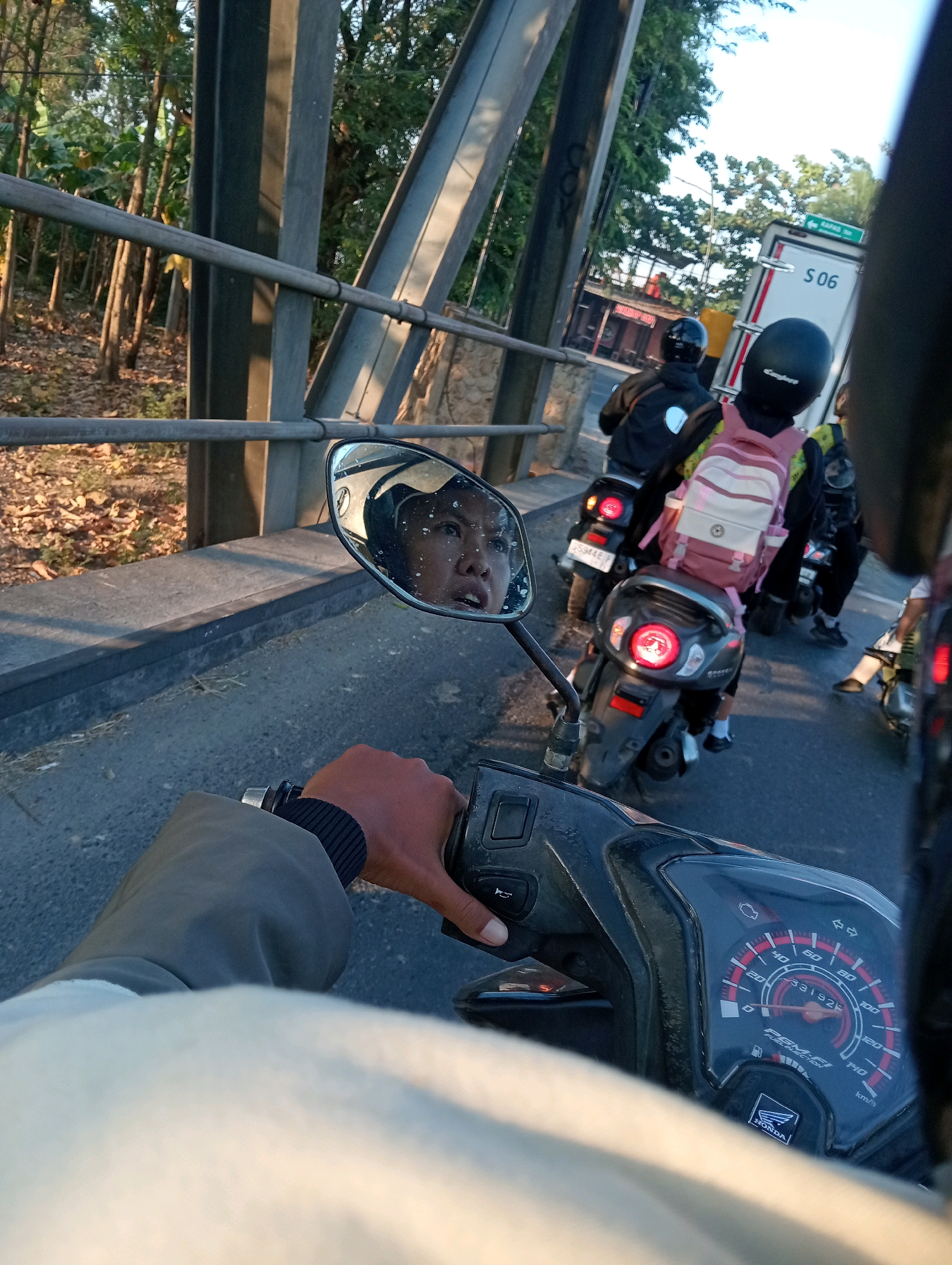 A first-person perspective of a person riding a motorcycle on a bridge in Bojonegoro, Indonesia. The rider's hand is visible on the handlebars, and the motorcycle's dashboard shows a speed of approximately 20 km/h. In the rearview mirror, the rider's face is reflected, showing a surprised expression. Other motorcycles and a white truck are also visible on the bridge, suggesting a traffic situation. The environment consists of a bridge structure, trees lining the roadside, and a clear sky, indicating daytime.