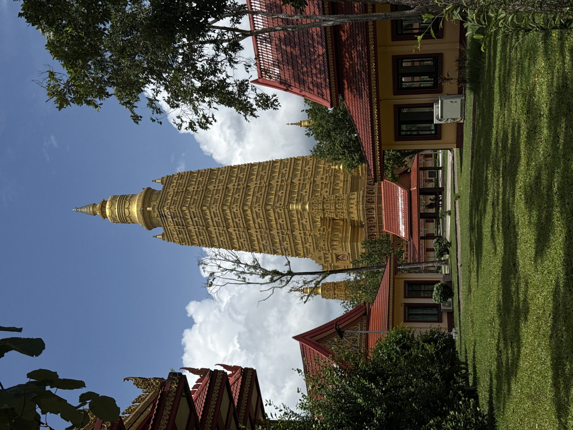 This image depicts the Wat Phra That Doi Suthep temple in the city of Na Nuea, Thailand, on a sunny day with a partially cloudy sky. The focal point is a towering, ornate golden stupa, characteristic of Thai Buddhist architecture, which dominates the background. In the foreground and midground, several other temple buildings with red tiled roofs and yellow walls are visible. Lush green grass covers the ground, with scattered trees and bushes adding to the natural environment. The overall scene conveys a sense of tranquility and spiritual importance.