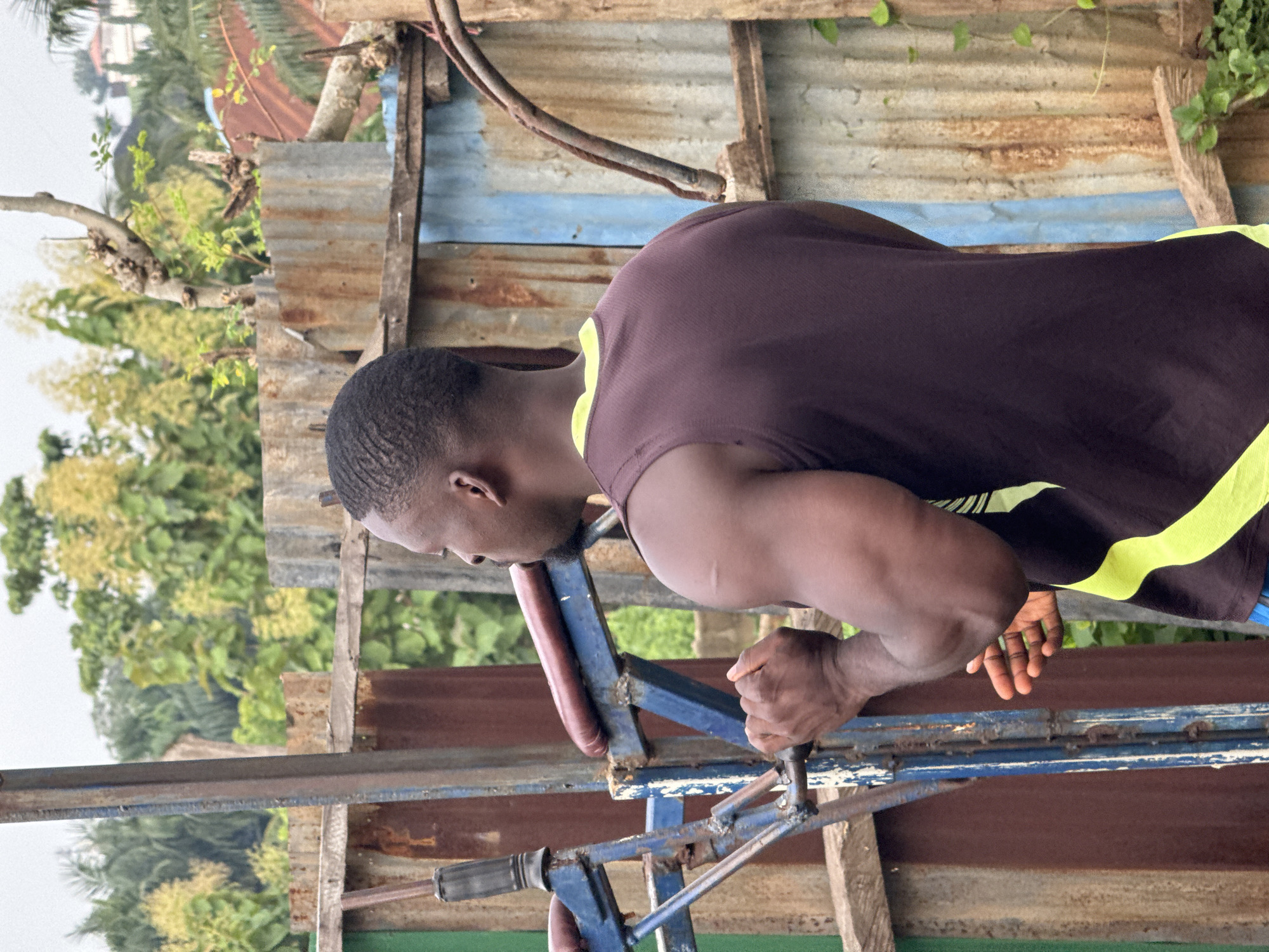 A Black male is pictured from the back, facing right, engaged in what appears to be a strength training exercise. He has short, dark, textured hair and is wearing a dark sleeveless top with a bright yellow trim around the neckline. His musculature is evident, particularly his deltoid and biceps. His hands are clasped around a blue metal bar, part of a piece of workout equipment. The equipment itself shows signs of wear and is mounted on a structure made of various corrugated metal sheets and wooden planks. The background consists of lush green foliage and trees, suggesting an outdoor setting. The overall environment appears to be somewhat makeshift, possibly a local gym or an outdoor workout area. The lighting suggests an overcast day. The location is identified as Akwanga, Nigeria.