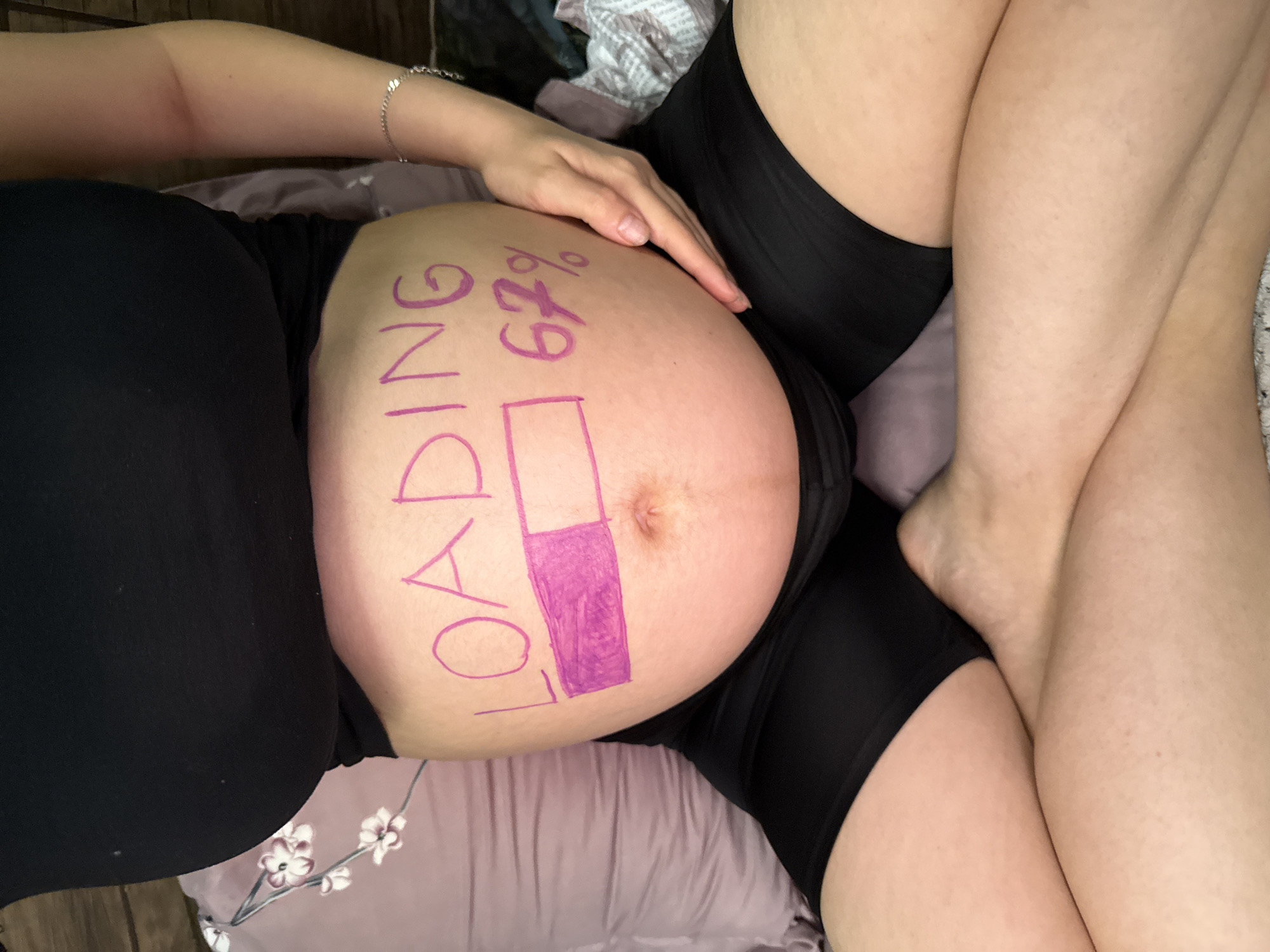 This is a top-down, close-up shot of a pregnant person's belly, filmed indoors. The person is lying down, wearing black clothing. The belly has "LOADING 67%" written on it in purple marker, with a progress bar indicating 67% completion. The person's hand is resting on their belly. The background consists of bedding and a wooden surface. There are no explicit indicators of time of day, weather, or strong emotions, but the text suggests an anticipation of birth. The provided location context is Tbilisi, Georgia.