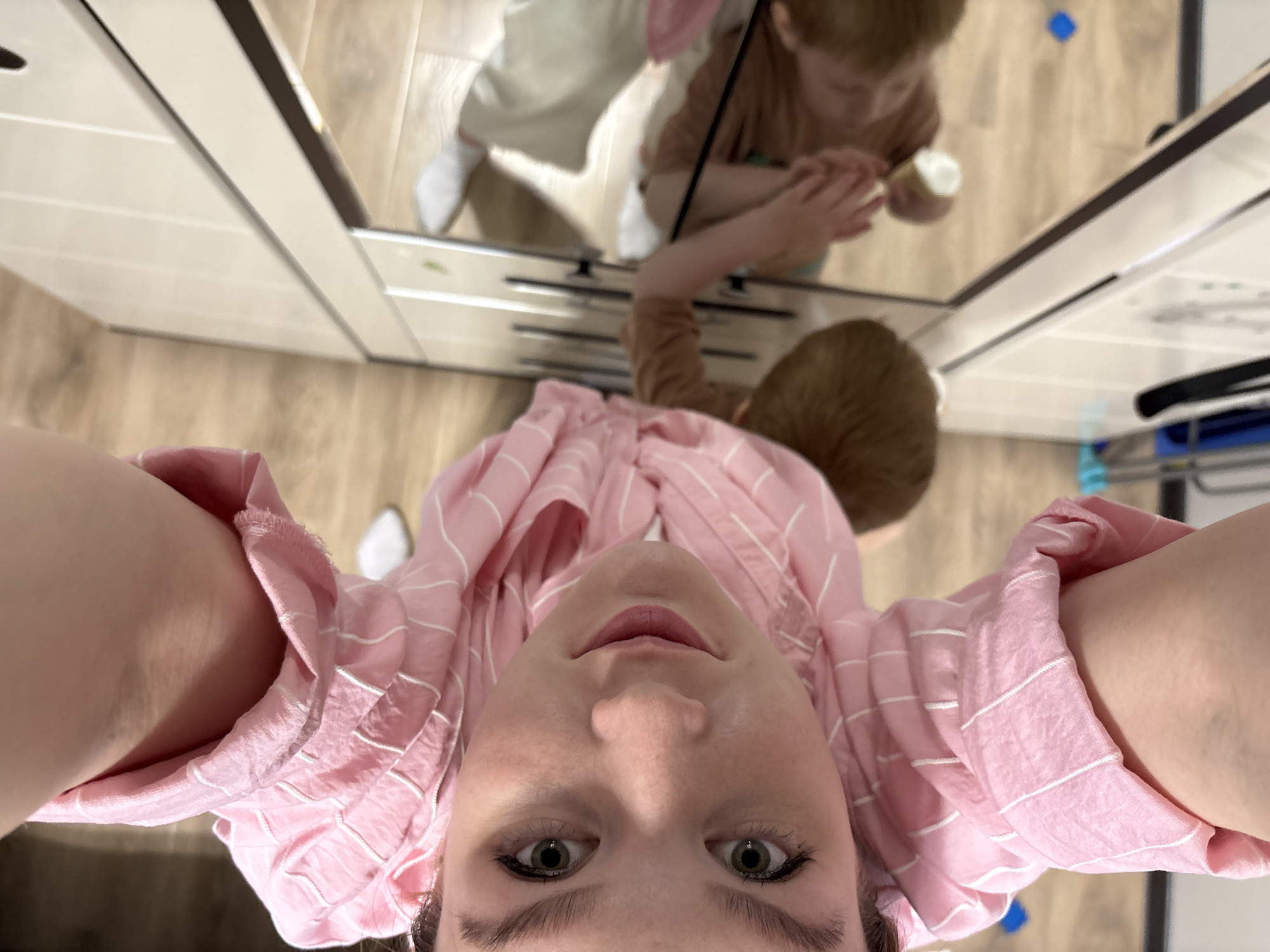 A high-angle selfie features a woman wearing a pink and white striped shirt. Above her head, a mirror reflects a child in a brown shirt seated and holding a white object. The room has light wood floors, light-colored cabinetry, and a blue object on the floor near the mirror's edge. The location in Izhevsk, Russia is indeterminate without further visual evidence.