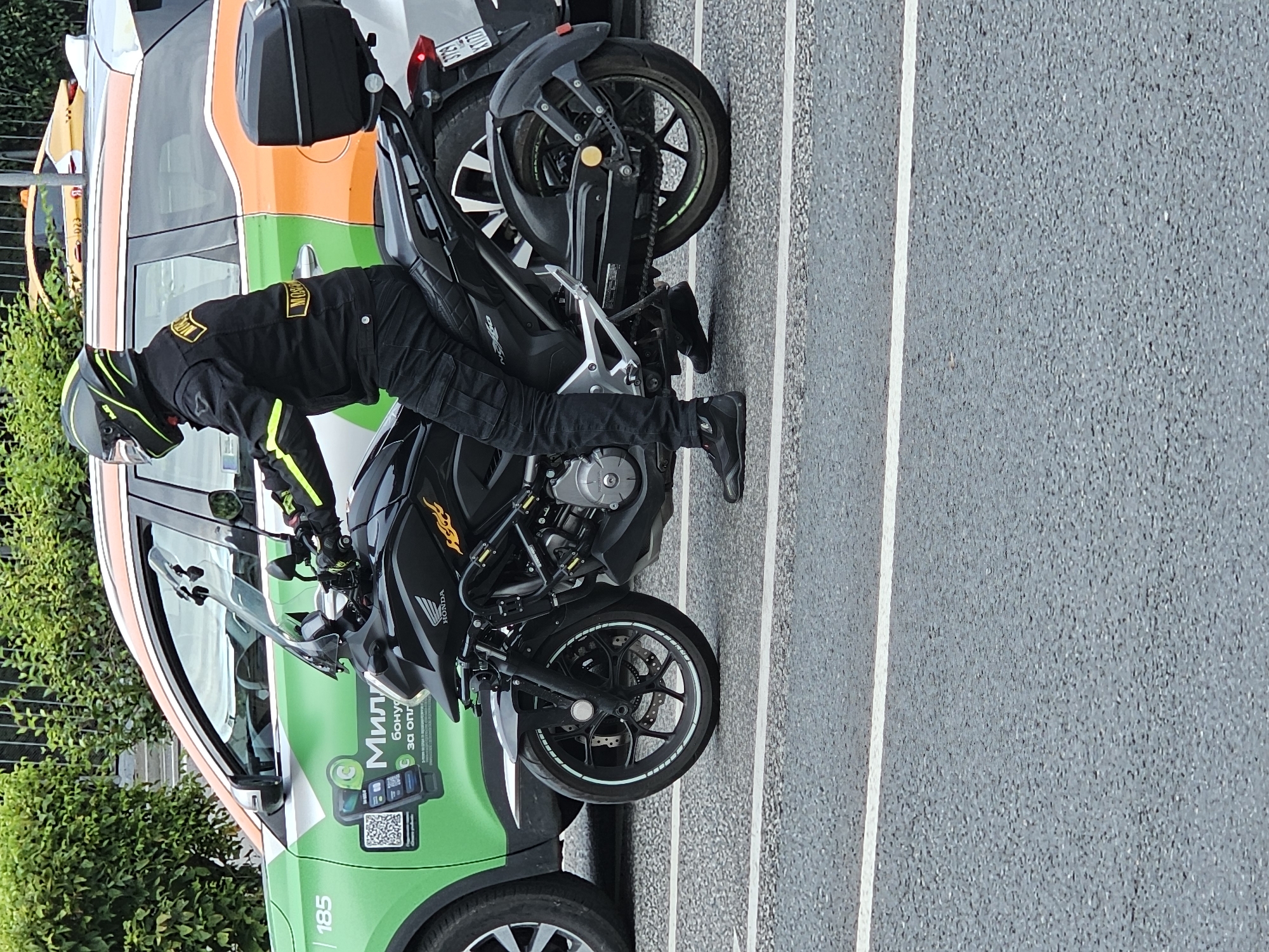 A man in full motorcycle gear, including a helmet, jacket, and pants, sits on a black Honda motorcycle. The motorcycle has a black top box and a tinted windshield. The man is wearing black gloves and black shoes. The motorcycle is in motion on a paved road, with white lines marking lanes. To the left of the motorcycle, a green and orange car is visible, with "185" printed on its side and Russian text that appears to be advertising a mobile service. In the background, a yellow taxi is also visible. The scene appears to be a street in the city of Khimki, Russia, given the context of the surrounding vehicles which are characteristic of Moscow region taxis and cars. The lighting suggests it is daytime.
