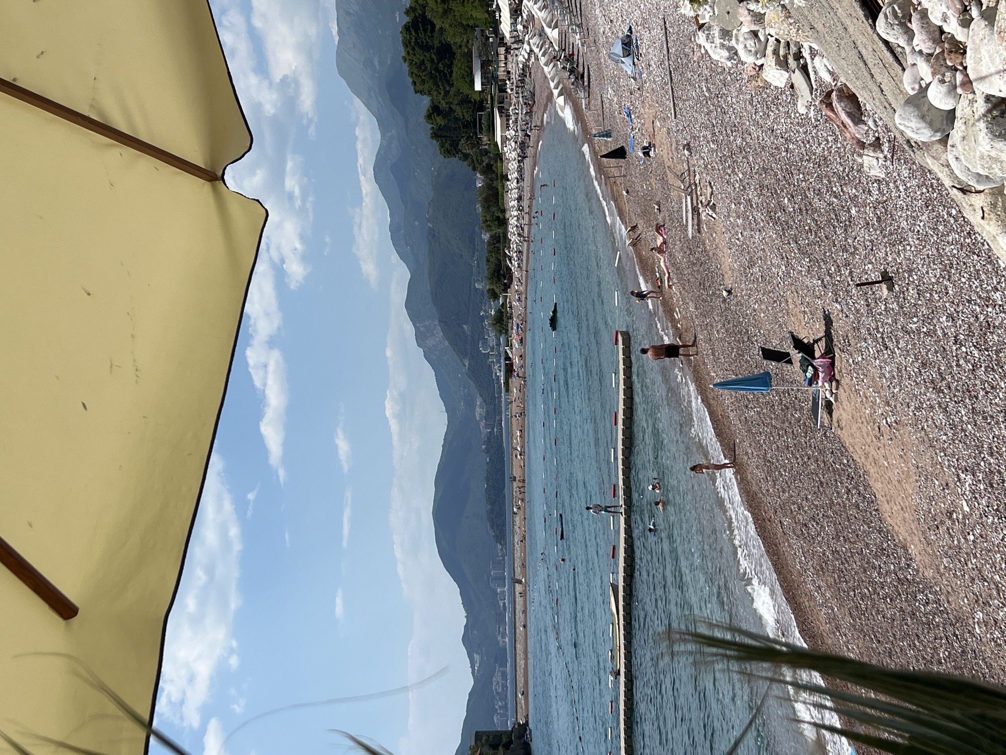 This media file captures a scenic view of a pebble beach in Sveti Stefan, Montenegro. The image is oriented vertically, with the bottom of the frame showing a segment of a light-colored beach umbrella, suggesting a shaded viewpoint.
The primary subjects are the beach, the sea, and the people present. A long stretch of pebble beach is visible, lined with numerous white beach chairs and umbrellas, indicating a popular tourist destination. The sea is a vibrant blue, with gentle waves lapping at the shore. In the water, several individuals are seen swimming and wading, enjoying the ocean. On the beach, a few people are sunbathing, walking, or interacting.
In the background, majestic green mountains rise, creating a dramatic backdrop for the coastal scene. Some buildings are visible nestled at the foot of the mountains, adding to the sense of a developed resort area. The sky is a clear blue with scattered white clouds, suggesting fair weather, likely during the daytime given the bright lighting and shadows.
A notable detail is a long wooden pier or barrier extending into the sea, marked with red and white buoys. This structure appears to delineate a swimming area. The overall mood is one of a relaxed summer day at the beach, with people enjoying the sun, sea, and mountainous scenery. There is no visible text in the image.