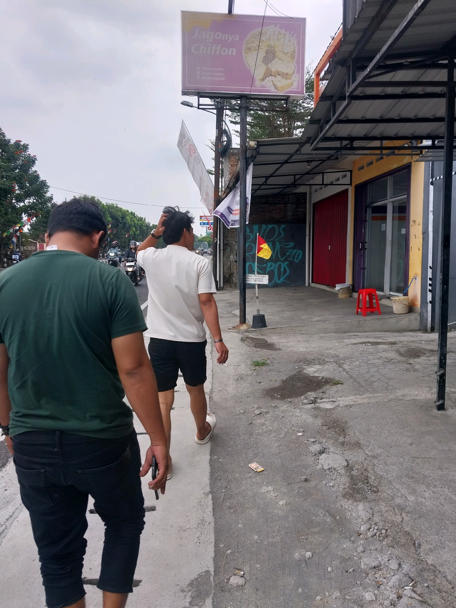 Two individuals are walking away from the camera on a paved sidewalk in front of a row of shops. The person in the foreground is wearing a dark green t-shirt and black cropped pants. The person behind them is wearing a white t-shirt and black shorts. They are walking on the sidewalk in the city of Sleman, Indonesia, as indicated by the signage visible in the background. The shops have metal awnings and various colored doors and windows. There is a red plastic stool and buckets outside one of the shops. The environment appears to be an urban street scene with trees lining the background.