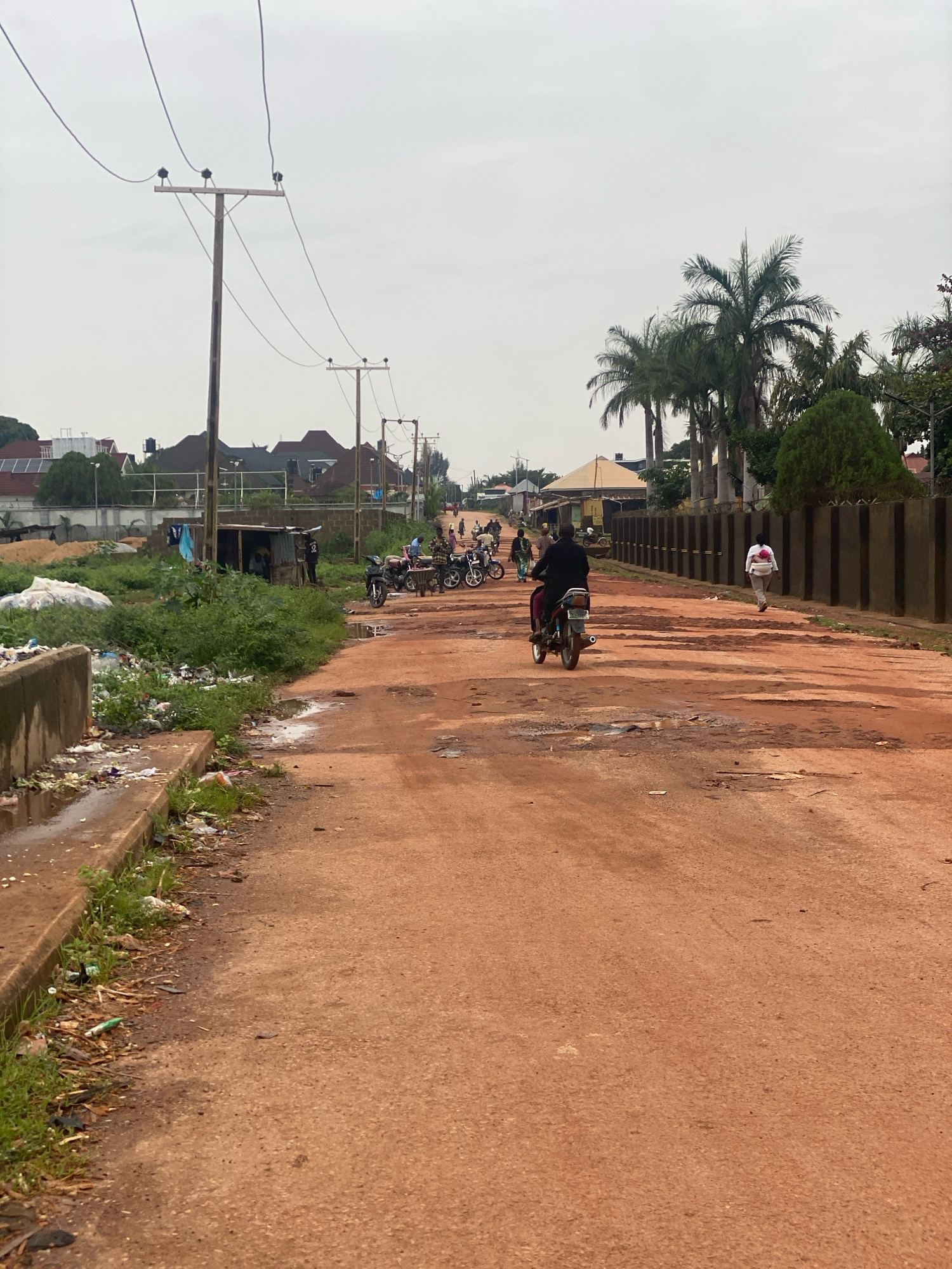 The image depicts a dusty, red dirt road in Narayi, Nigeria, with visible potholes and uneven surfaces. The road is lined with sparse vegetation and a concrete barrier on the left, which is strewn with garbage. Several people are present, some walking and others on motorcycles. In the foreground, a person is riding a motorcycle away from the camera. Further down the road, a group of motorcycles is parked, and people are gathered around a small roadside stall. Buildings and palm trees are visible in the background, suggesting a suburban or semi-urban environment. The sky is overcast. Utility poles with electrical wires are also prominent features in the scene.