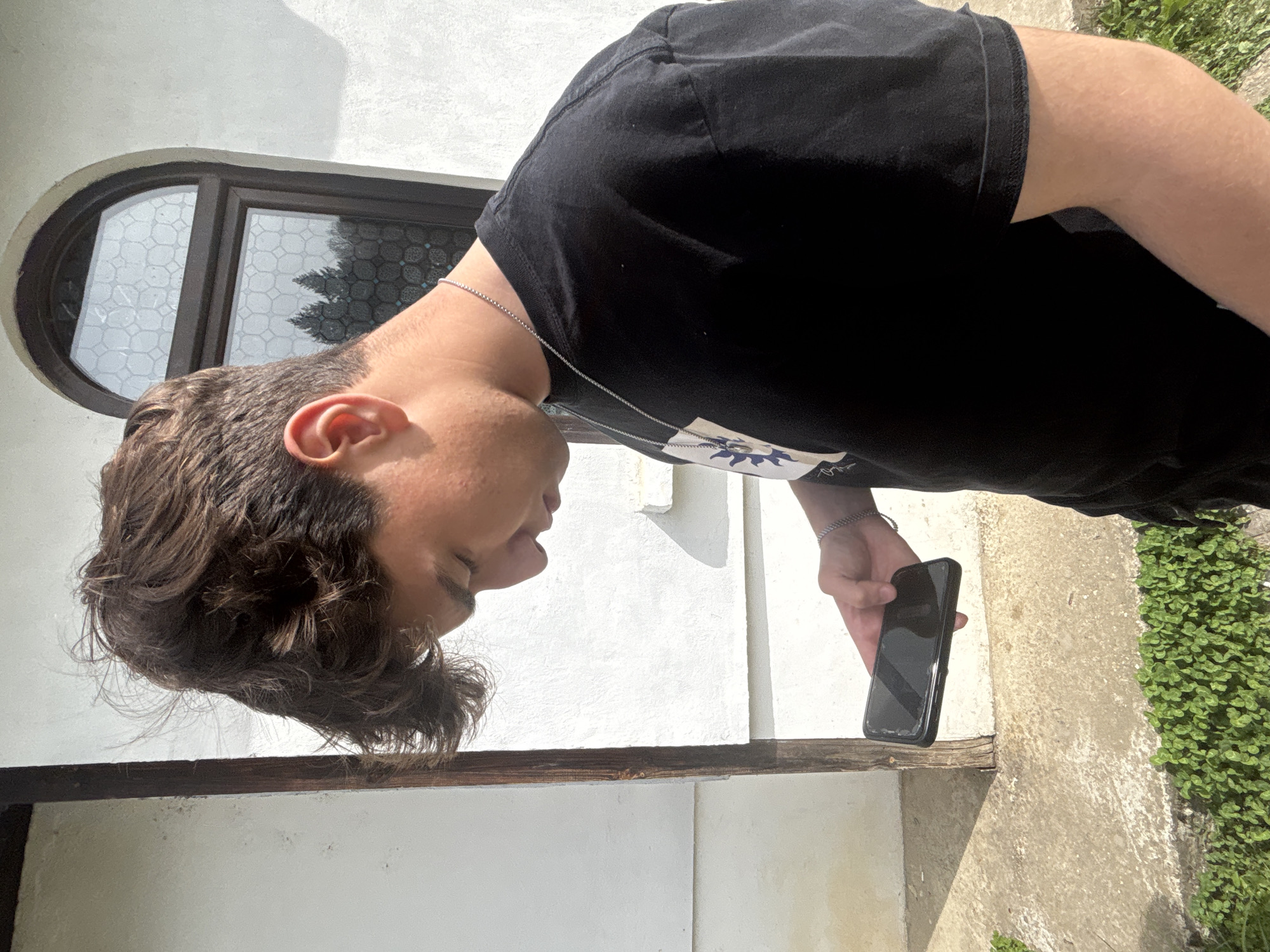 A young man stands outside, holding a smartphone in his left hand. He has dark hair, a light complexion, and wears a black t-shirt and a silver chain. He is standing next to a white wall with a unique, horizontally-oriented oval window with patterned glass. Ground cover is visible to the right. The location is Troyan, Bulgaria.