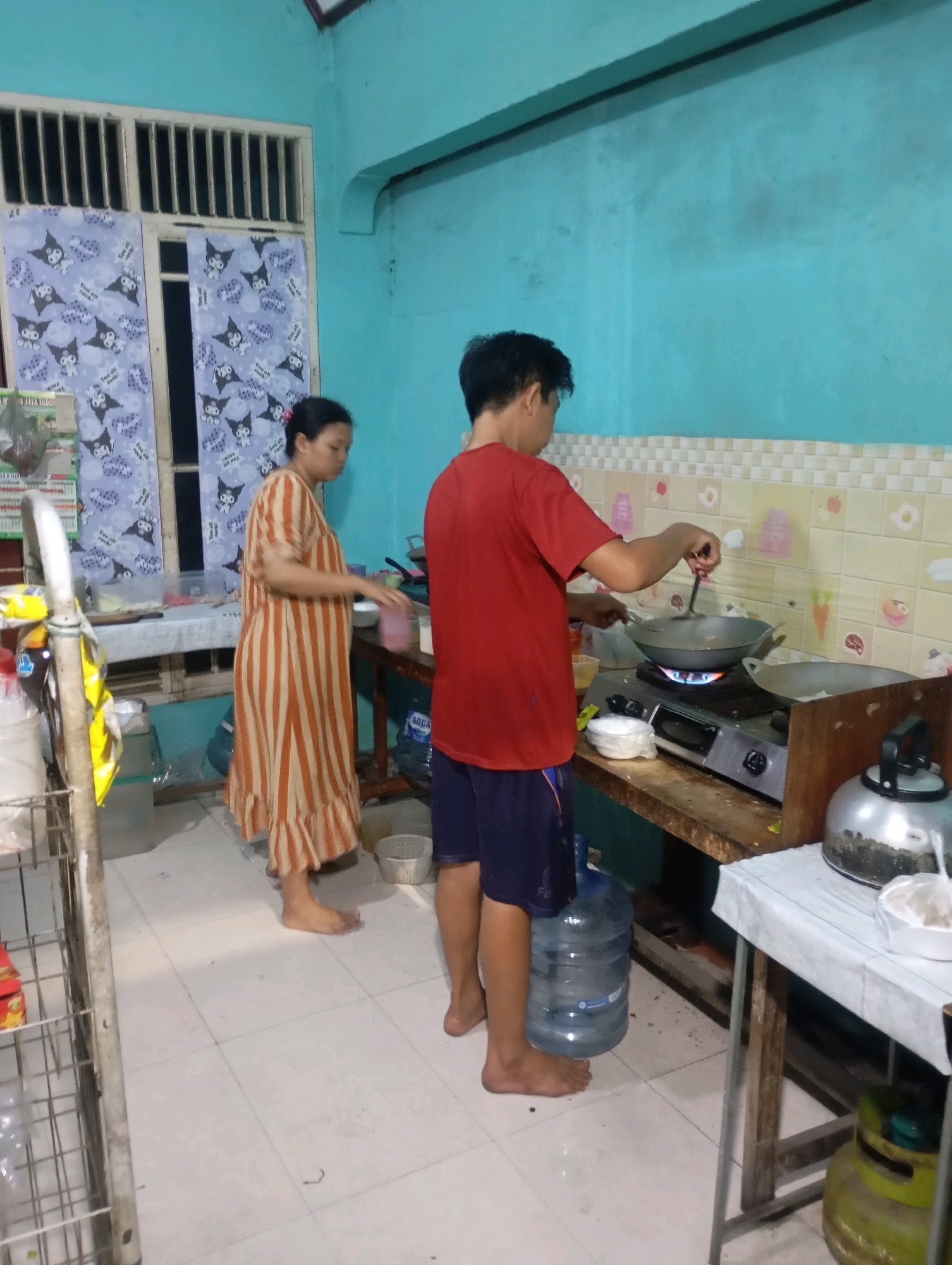 In Babelan, Indonesia, a young man wearing a red t-shirt and dark shorts is cooking in a kitchen. He is standing in front of a gas stove, stirring food in a wok. A woman in a striped dress stands nearby, facing away from the camera. The kitchen has light blue walls, a tiled backsplash, and a wooden countertop. There are various kitchen items visible, including pots, a kettle, and a large water jug. The floor is tiled with white squares.
