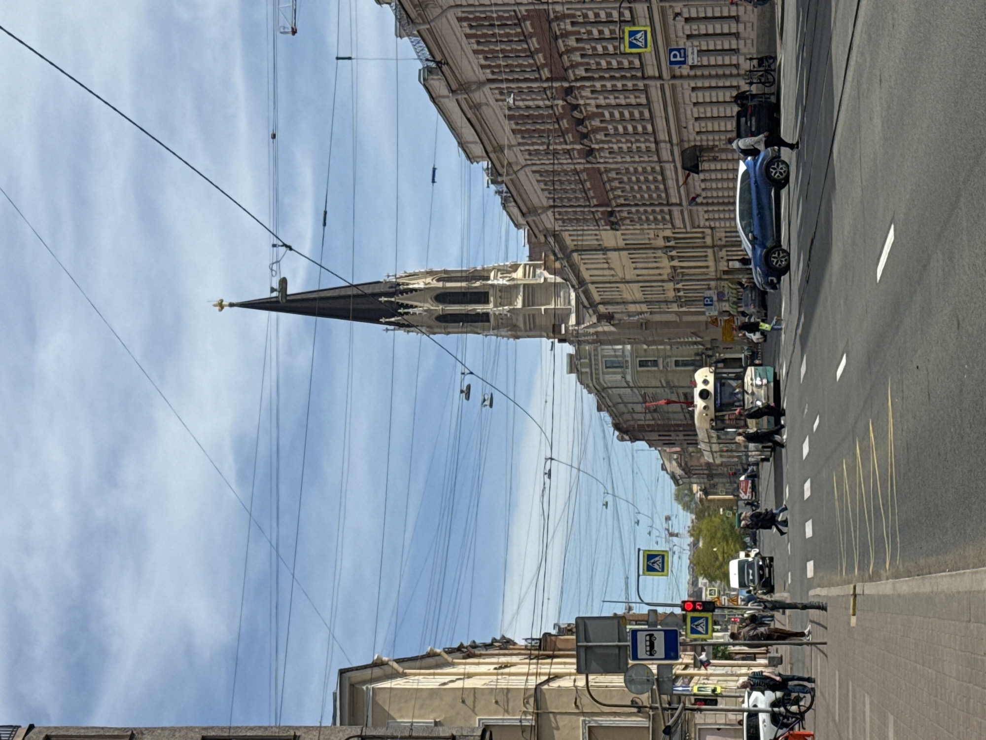 The image depicts a street scene in Saint Petersburg, Russia, dominated by a tall, gothic-style spire of the Church of Saint Anne. Overhead electric wires crisscross the sky. On the right side of the street, there is a blue car parked on the roadway. Pedestrians are seen walking on the sidewalk and crossing the street. There are traffic signals and road signs visible. Buildings with detailed facades line the street. The weather appears to be sunny with some clouds in the sky.