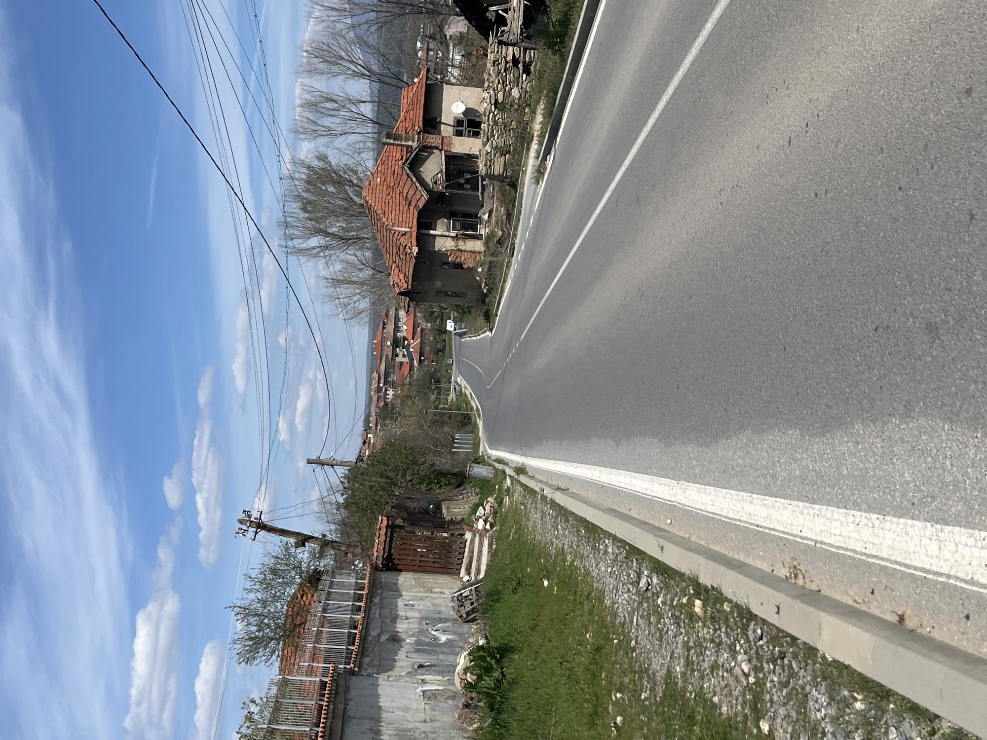 The image depicts an empty asphalt road running through a rural or suburban area. The road has double white lines marking the lanes. On either side of the road are buildings and houses, some of which appear old and dilapidated, with damaged or missing roofs. There are also fences, grass, and gravel along the roadside. Overhead, there are power lines stretching across the sky, which is blue with scattered white clouds. The buildings suggest a small town or village setting.
