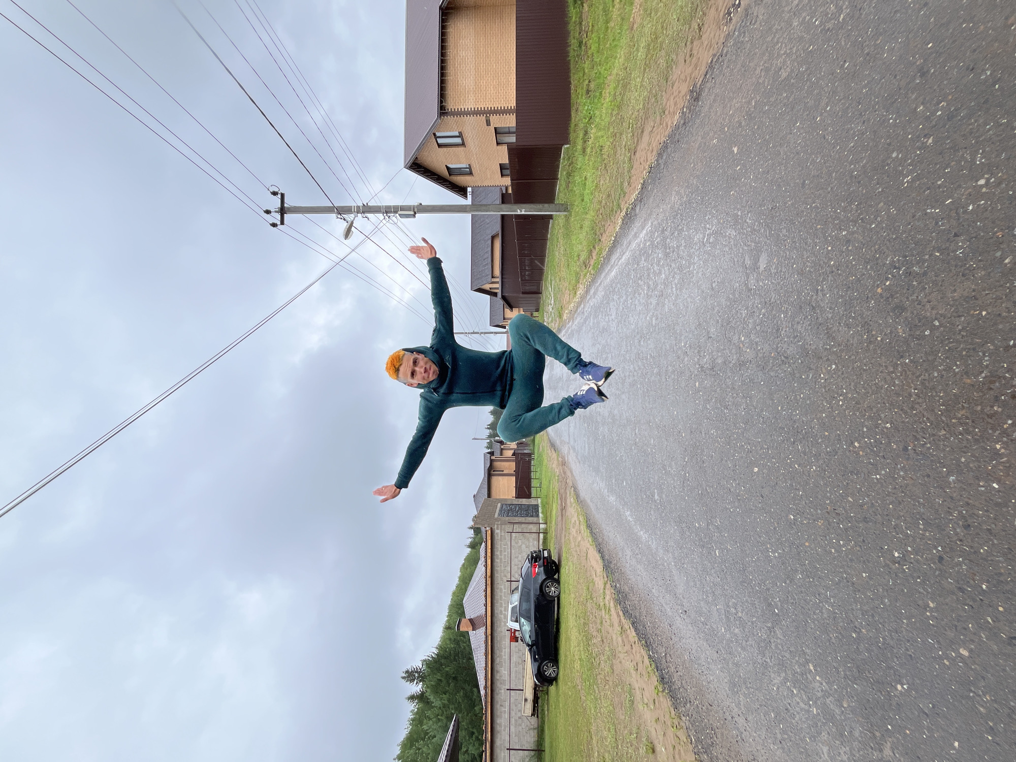 A young man with bright orange hair is captured mid-air, mid-jump, on a wet, asphalt road. He is wearing a dark green tracksuit and blue sneakers. His arms are outstretched to the sides, with his palms facing forward, and his body is positioned horizontally. He appears to be in a suburban area, indicated by the presence of houses and fences on either side of the road. In the background, a black car is parked on the left, and houses made of brick are visible on the right, some with brown fences. A utility pole with multiple wires stretches across the frame, contributing to the overcast, cloudy sky. The overall scene suggests a slightly damp, cool day in a residential area of Chaikovsky, Russia.
