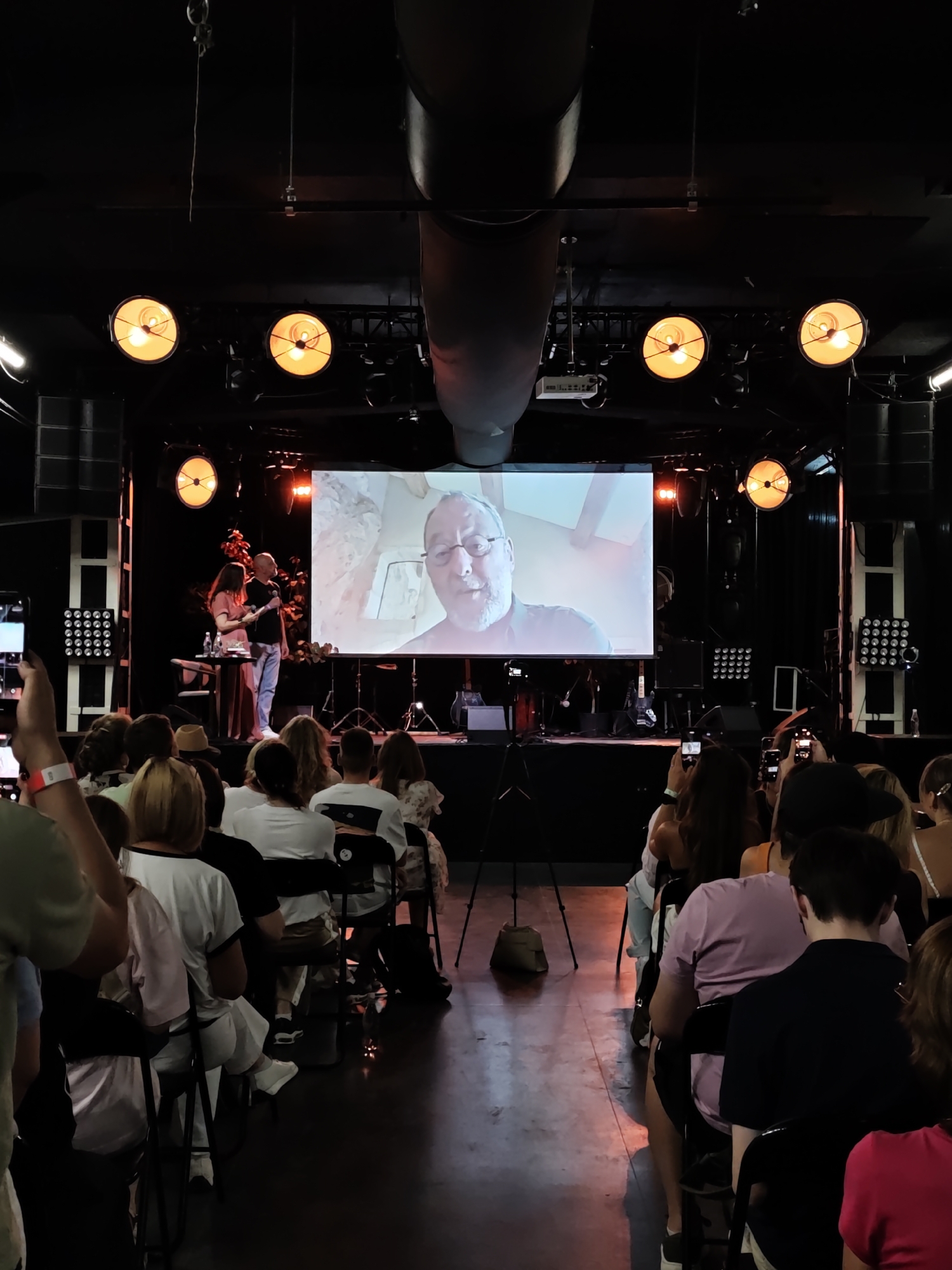 This image depicts an indoor event, possibly a presentation or a small concert, taking place in Lviv, Ukraine. The scene is viewed from the perspective of the audience, facing a stage area.
The stage features a large projection screen displaying a close-up of a man with glasses and a beard, seemingly engaged in a conversation or presentation. To the left of the screen, two individuals are standing on the stage: a woman in a long pink dress and a man in a light blue shirt and pants, holding a microphone. Behind them, a band or musical setup is partially visible.
Above the stage, several large, industrial-style spotlights with orange light illuminate the area. The ceiling appears to be dark, with visible pipes or ventilation systems. On the sides of the stage, large speaker systems are mounted.
The audience is seated in rows of chairs, facing the stage. Many individuals are holding up their mobile phones, presumably to record or photograph the event. The lighting in the audience area is dim, with the stage lights casting a warm glow on the floor. The overall environment suggests a contemporary event space.