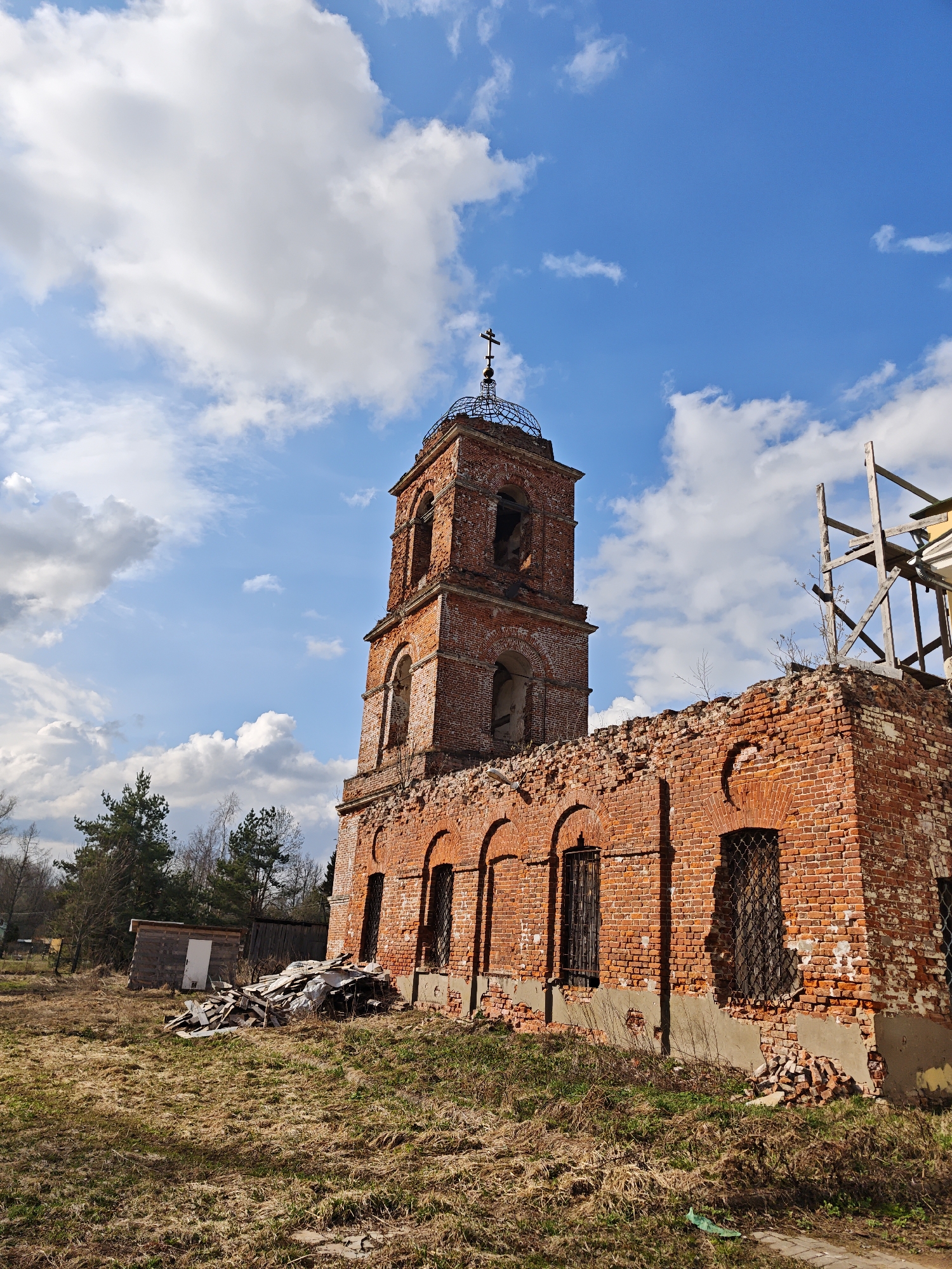 The image depicts the ruins of a red brick church in Ивановское (большерогачевский С/о), Russia, set against a bright blue sky with scattered white clouds. The church structure is heavily damaged, with missing sections of walls and the bell tower. The bell tower is capped with a metal dome and cross. Scaffolding is visible on a partially collapsed section of the church, indicating possible restoration efforts. The foreground shows an unkempt grassy area with debris. A small wooden outbuilding is visible in the background to the left.