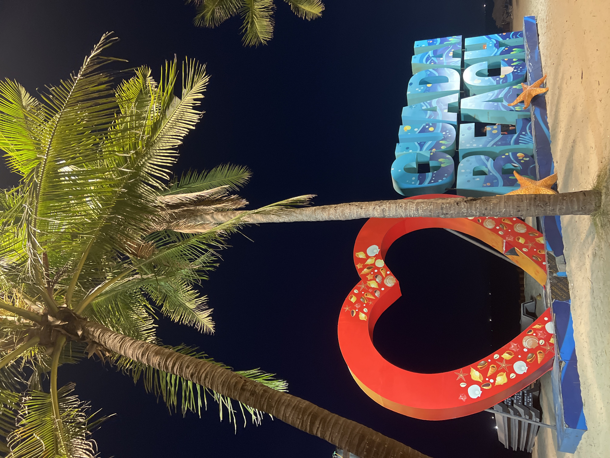 This is a nighttime, low-angle, eye-level shot of a beach attraction in Hoi An, Vietnam. The foreground features a large, red, heart-shaped sculpture adorned with shells and starfish. To the right of the heart is a large, blue, three-dimensional sign that reads "GUADA BEACH," with the letters decorated with ocean-themed imagery. Two large orange starfish figures are placed at the base of the sign. Two palm trees with illuminated green fronds are prominently featured in the left and right portions of the frame, with their trunks extending downwards. The background is a dark, clear night sky. The ground is covered in light-colored sand. In the middle distance, behind the heart sculpture, a stack of what appear to be concrete slabs is visible. Further back, a person is visible inside the "BEACH" section of the sign. The overall scene depicts a decorated tourist spot on a beach at night.