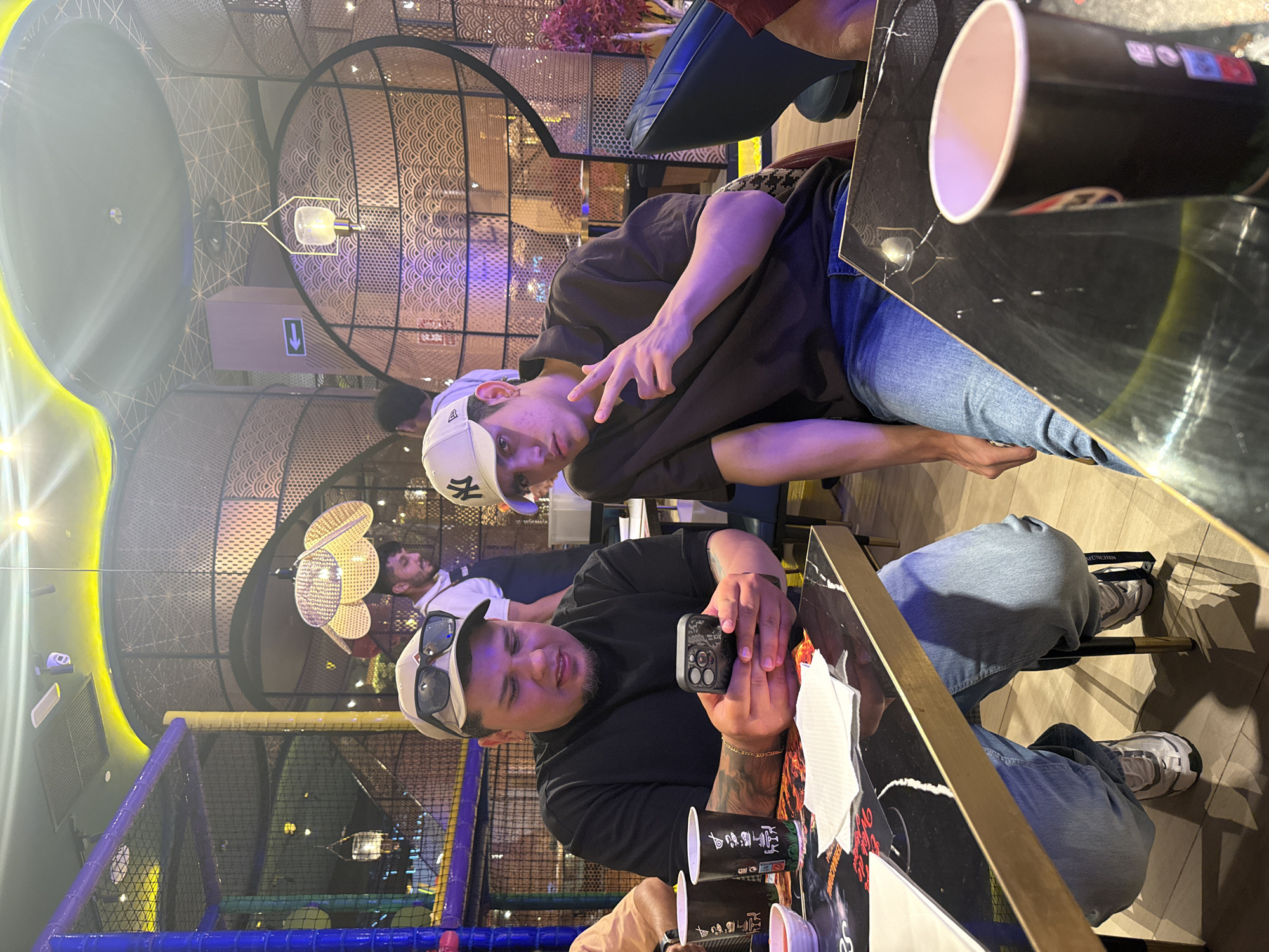 This media file captures two young men indoors, possibly at a restaurant or cafe, given the presence of tables, cups, and decorative lighting. One man, wearing a white baseball cap and a dark t-shirt, is making a peace sign with his fingers while looking at the camera. The other man, also wearing a cap and a black t-shirt, is holding a smartphone and looking towards the camera with a slight smile. They are seated at a table with a black, reflective surface, on which there are several cups and what appears to be a placemat with some text and graphics. The background reveals a modern interior with intricate latticework on the ceiling and walls, and some colorful lighting, suggesting a contemporary establishment. The overall mood appears casual and friendly, with the subjects looking relaxed and engaged with the camera. There is no indication of the time of day or weather conditions. The provided location context is Carretera De San Martín, Spain. Visible text includes "NY" on one of the caps and possibly branding on the cups.